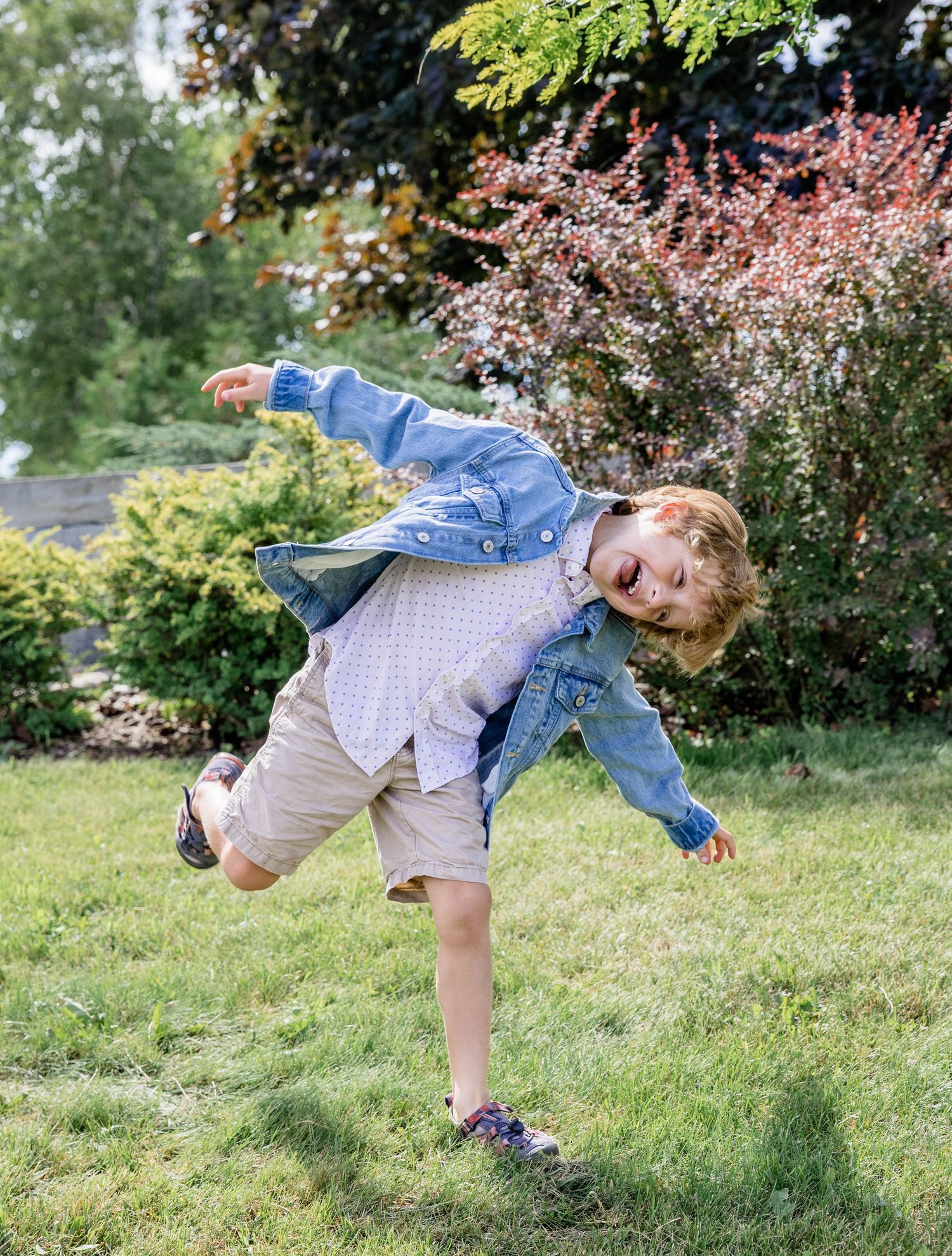 A young boy in a denim jacket and khaki shorts joyfully jumping and playing on a green lawn outdoors.