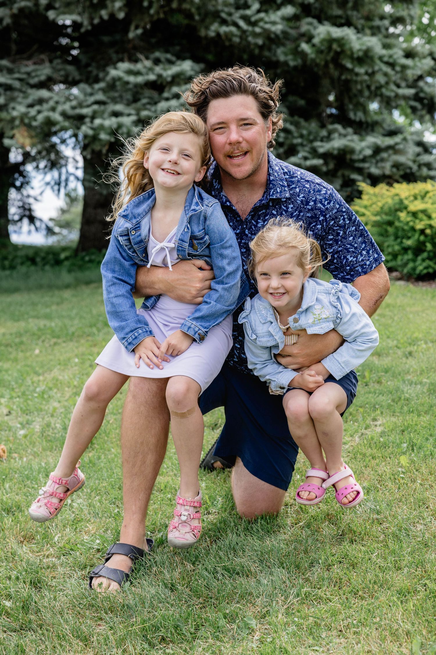 A man kneeling on green grass smiling with two young blonde girls in denim jackets outdoors near trees.