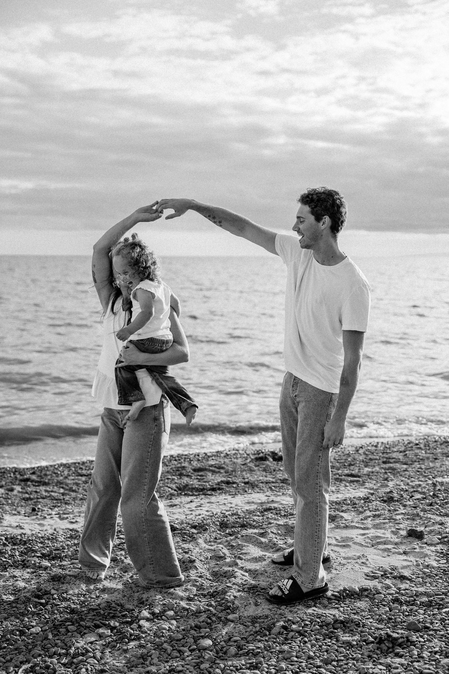 Black and white photo of a couple dancing on a pebble beach, man spinning woman near the ocean shore.