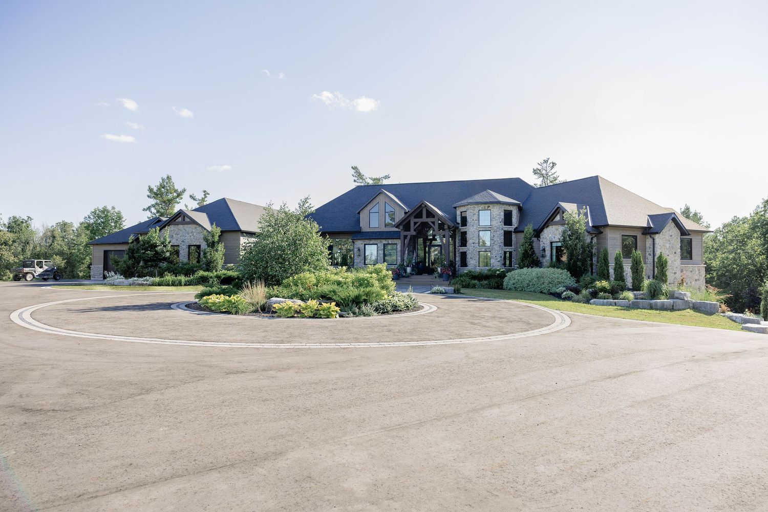 Grand luxury estate with circular driveway, manicured landscaping, and stone facade under blue sky.
