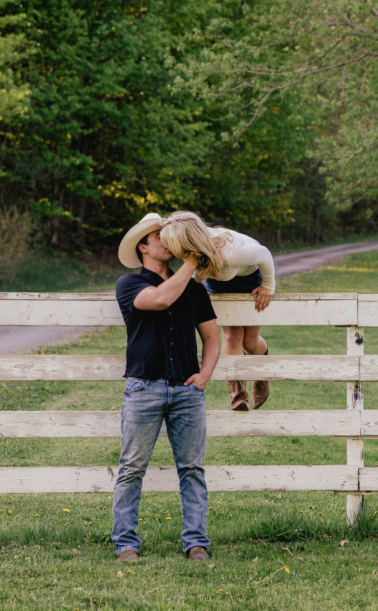 A cowboy in a white hat kisses a blonde woman sitting on a white wooden fence in a green field.