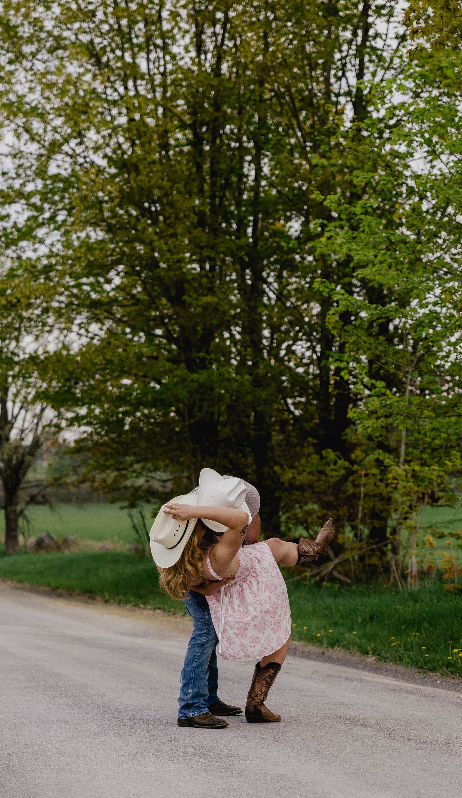 A couple shares a sweet kiss on a rural road, the woman in a pink dress and cowboy boots.