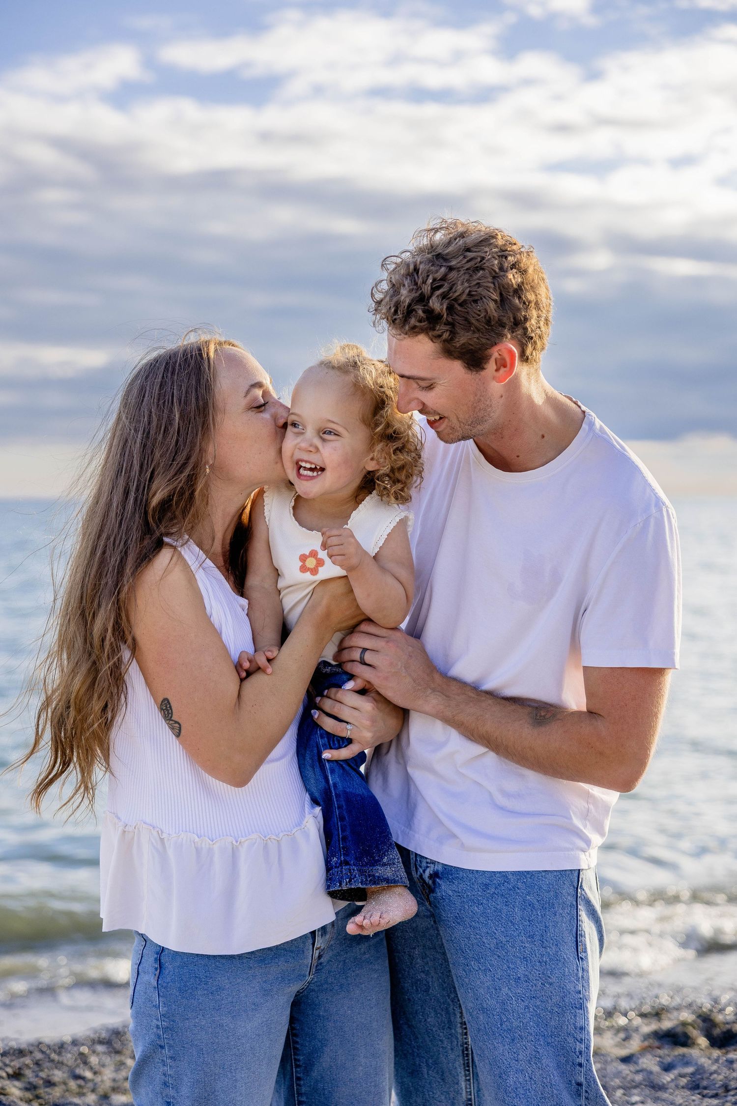 Happy young family with curly-haired toddler sharing a joyful moment on a rocky beach under cloudy skies.