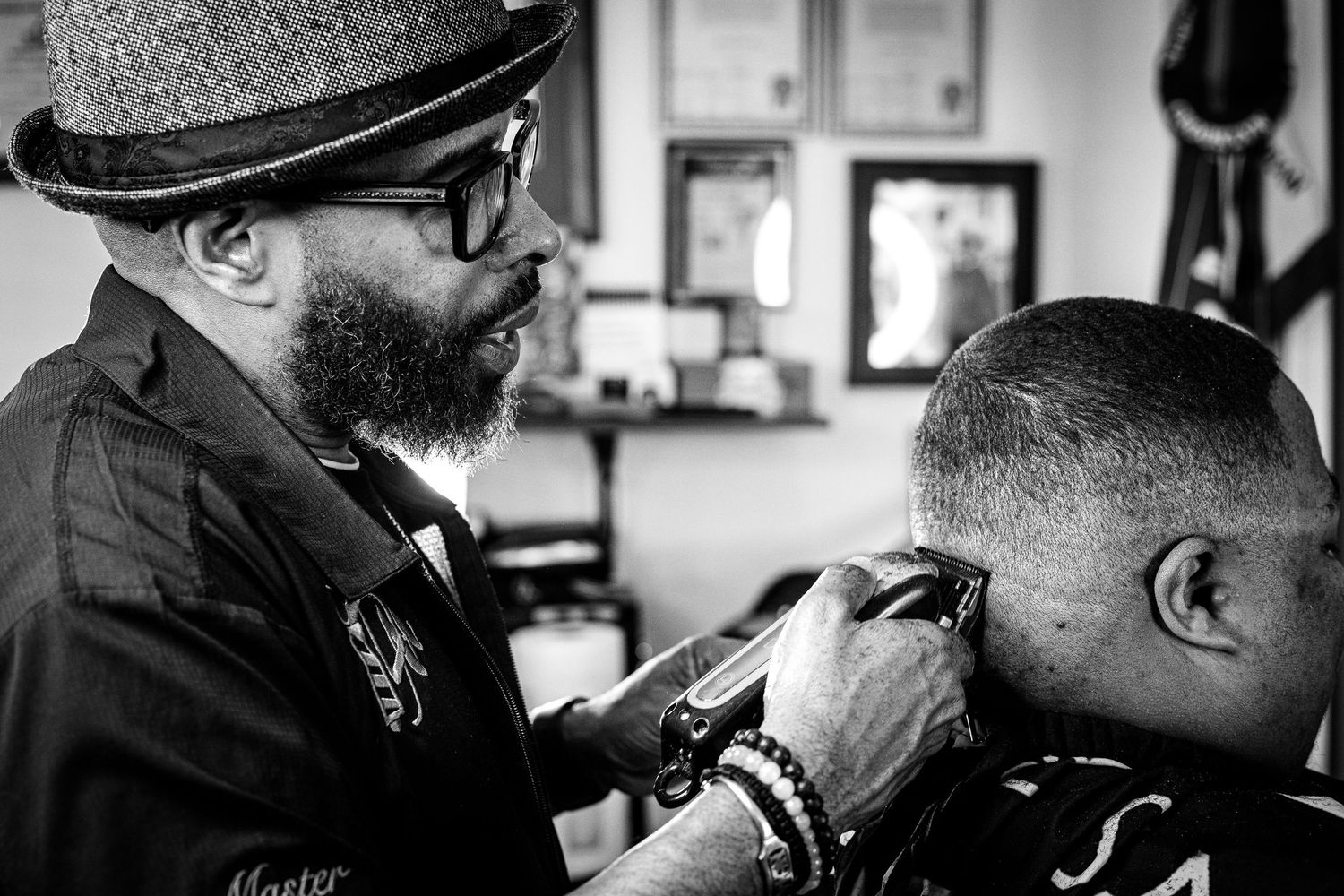 Black and white photo of a bearded barber in a hat carefully cutting a client's hair.