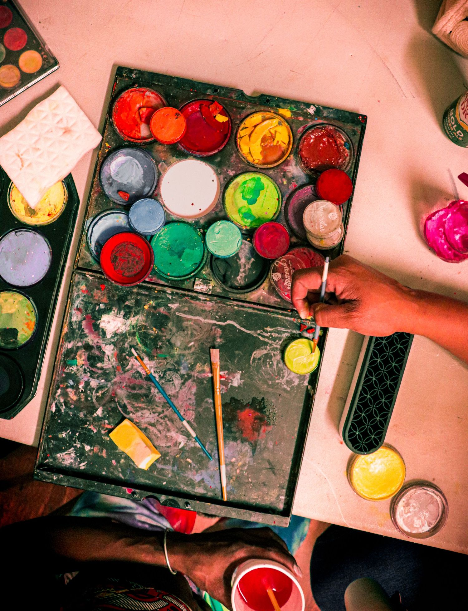 Overhead view of a vibrant face paint palette with a hand holding a brush mixing colors.