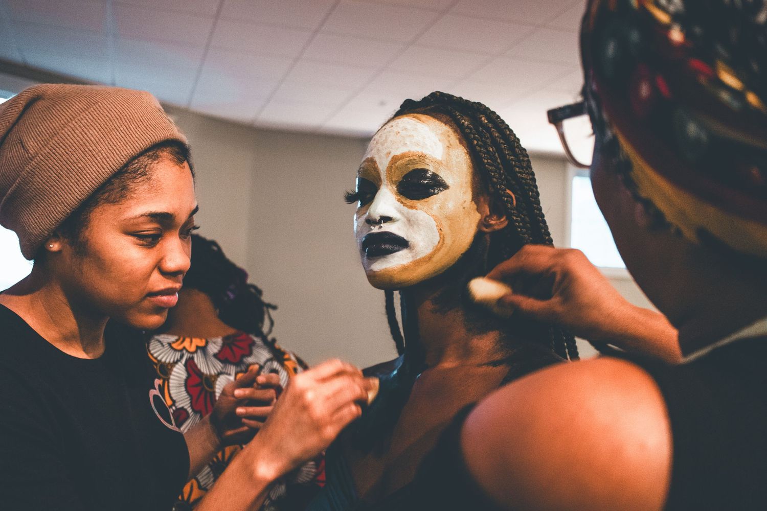 Two artists carefully apply white theatrical face paint to a performer with braided hair backstage.