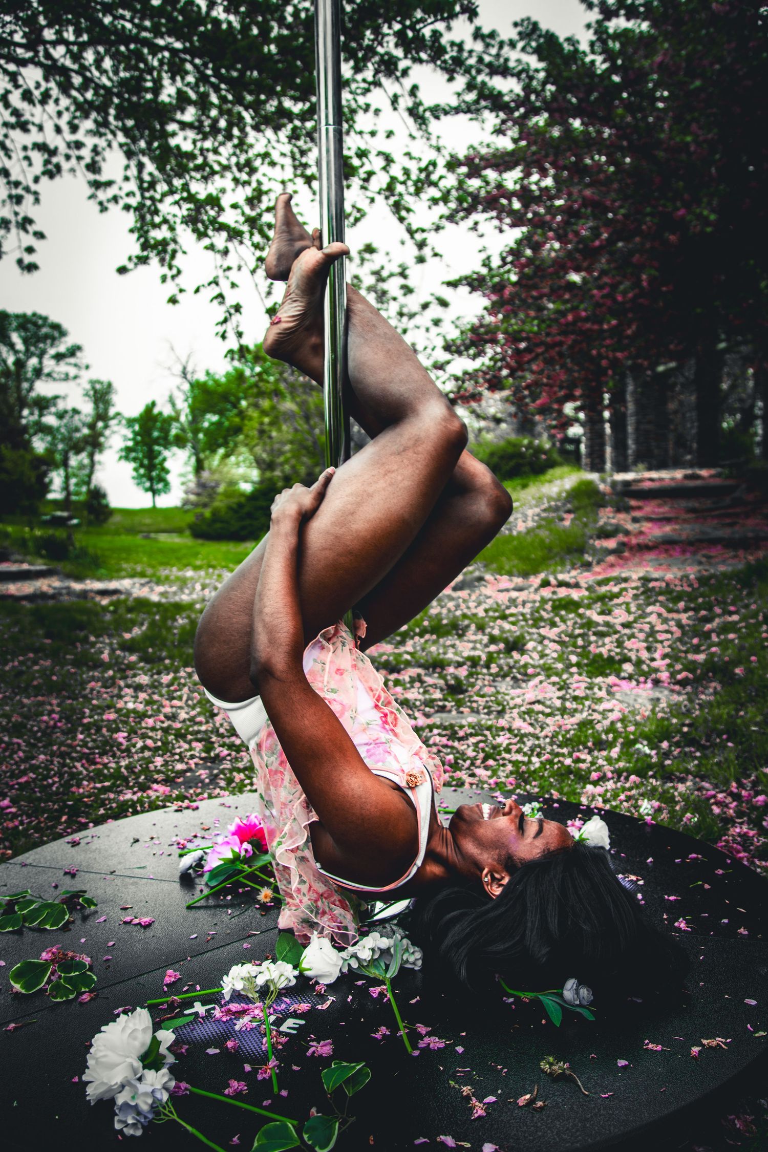 Woman performing inverted pole dance move outdoors surrounded by scattered flowers.