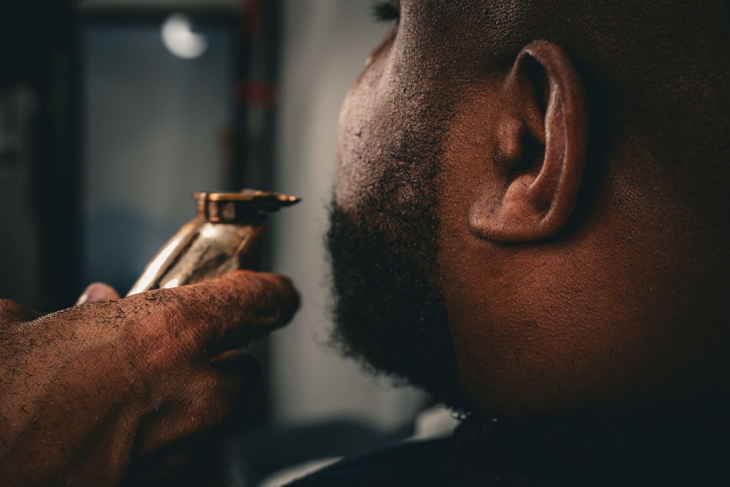 Close-up of a barber trimming a Black man's beard with gold clippers.