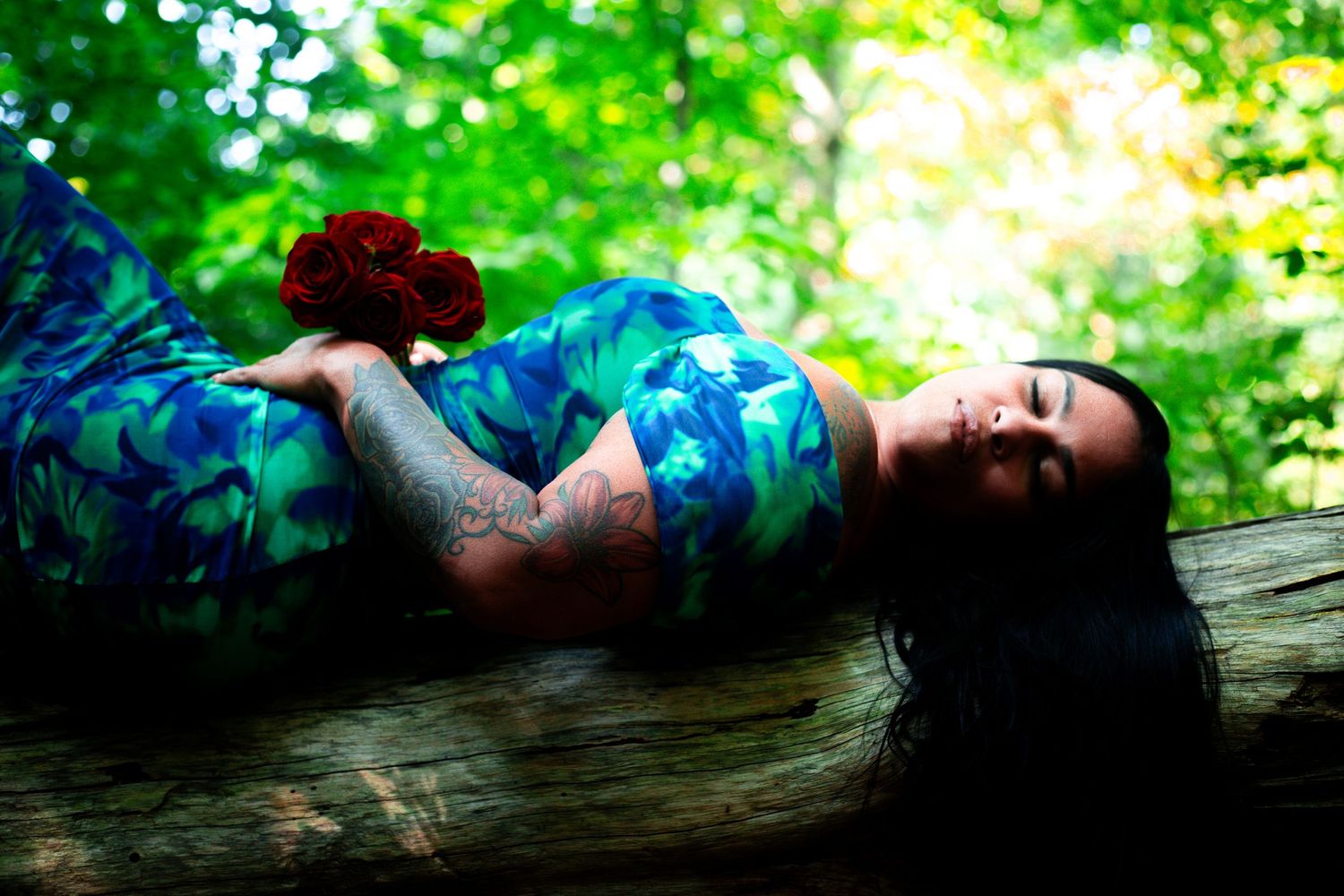 Woman in a blue floral dress lying on a fallen log in a lush green forest holding a red rose.