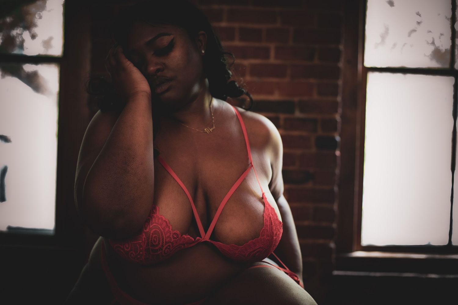 Curvy woman in red lace bralette seated pensively near a window with soft natural light and brick backdrop.