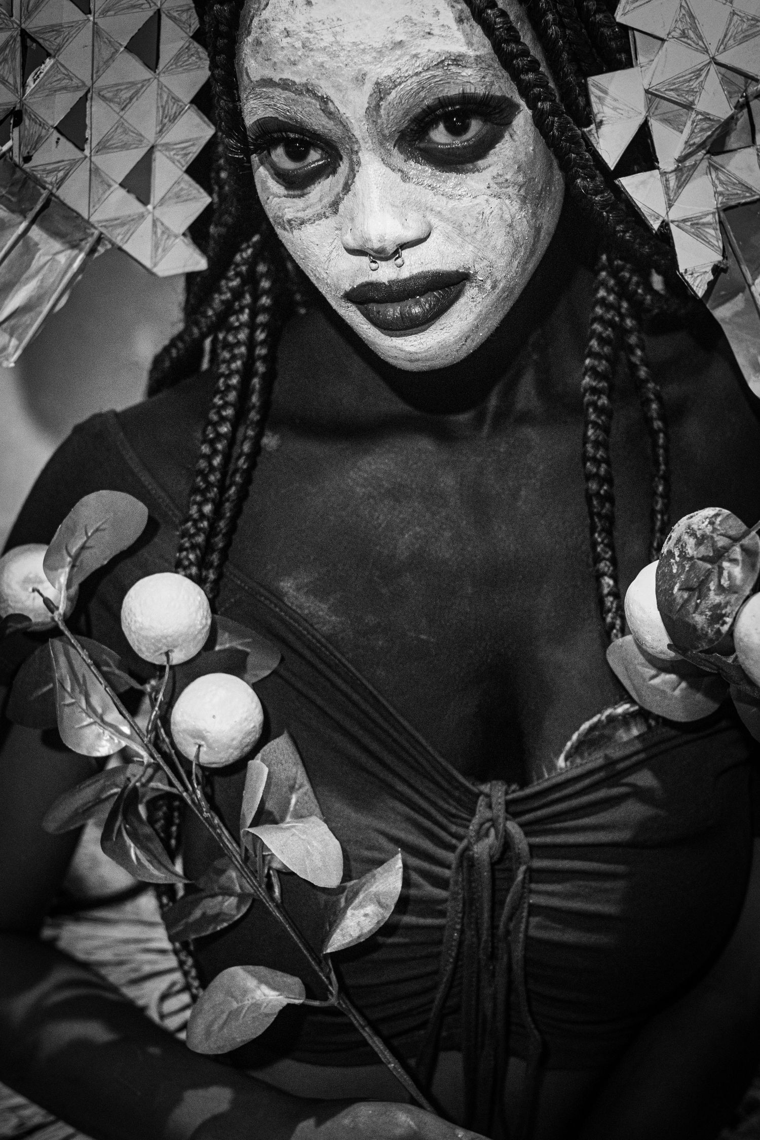 Black and white portrait of woman with white face paint, braids, and nose ring holding a floral branch.