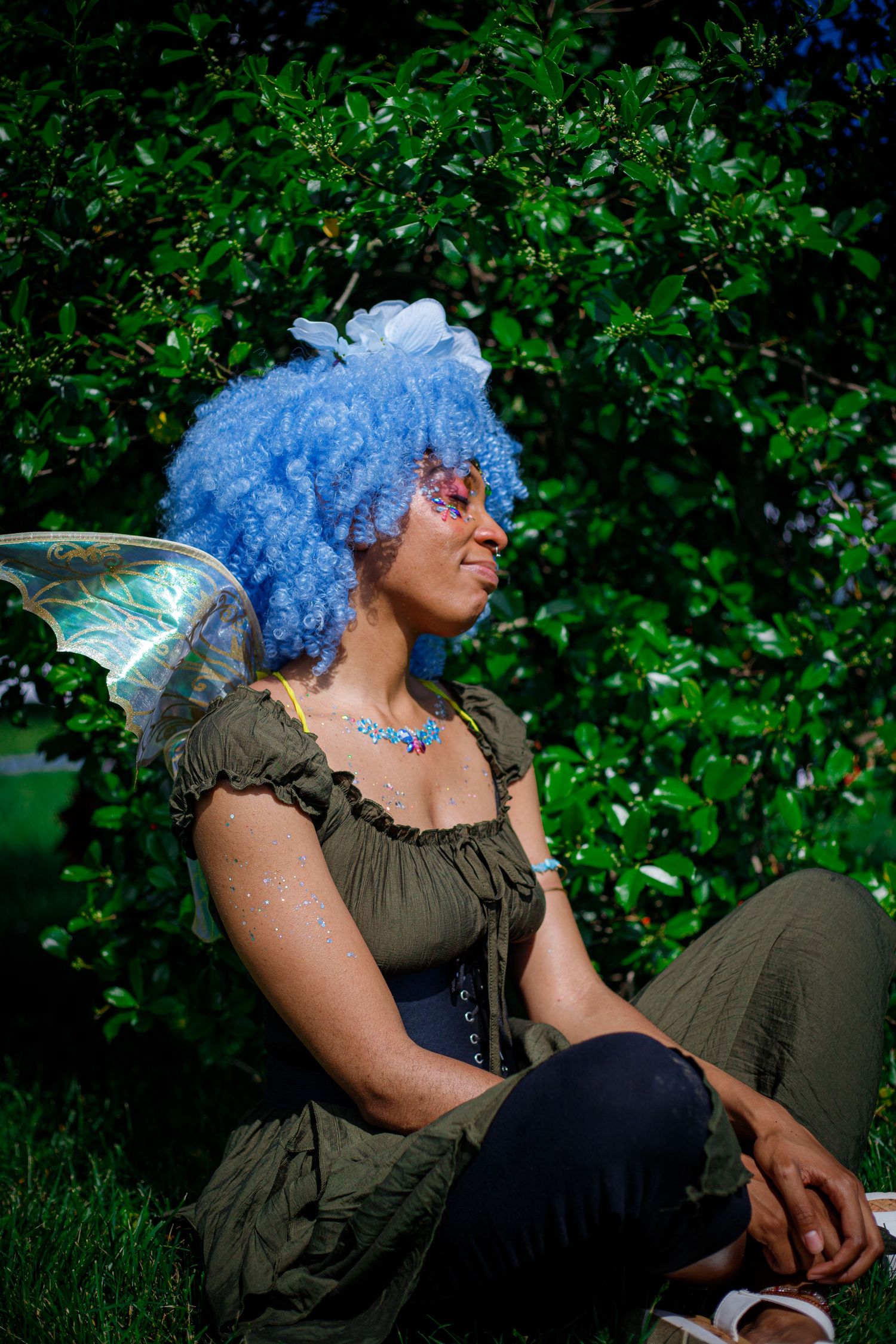 Woman in blue curly wig and fairy wings sits outdoors in green nature setting wearing fantasy costume.