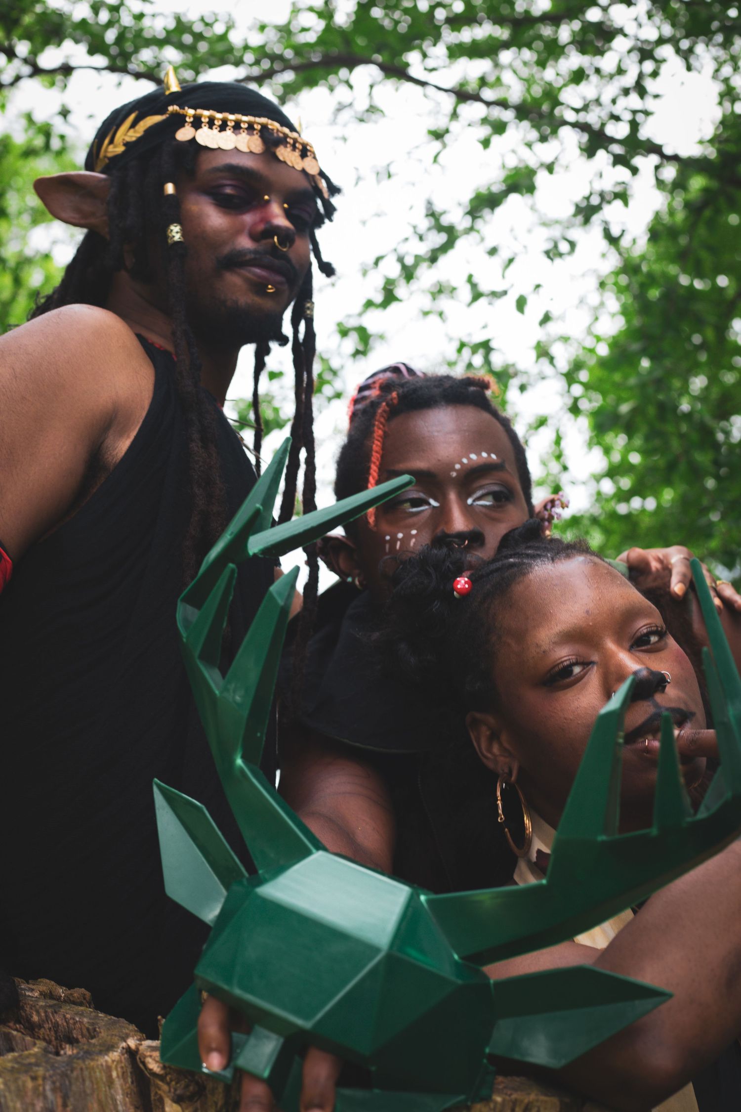 Three people in fantasy elf costumes holding green geometric prop pose together among forest trees.