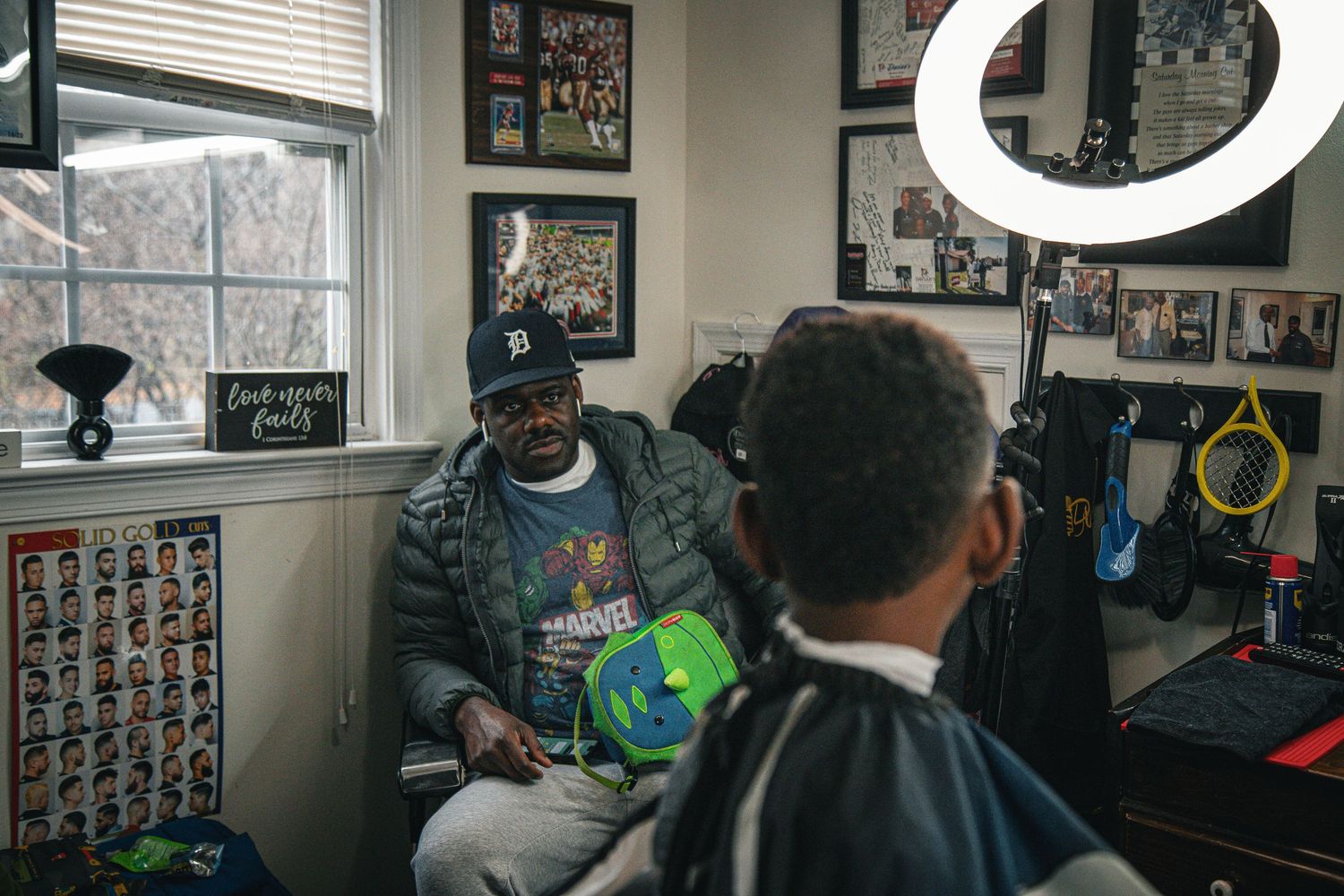 A barber consults with a young client in a decorated barbershop with ring light and framed photos.