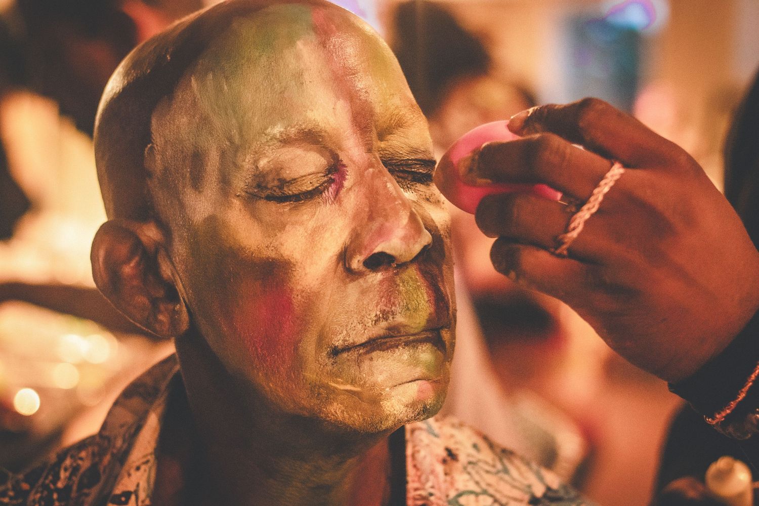 Close-up of green and red theatrical face paint being carefully applied to an older mans face.