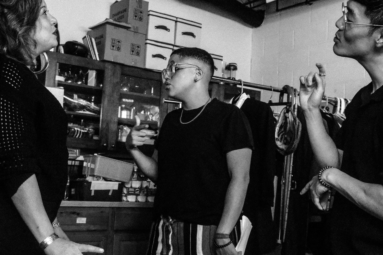 Black and white photo of three people having a conversation in a cluttered backstage storage room.