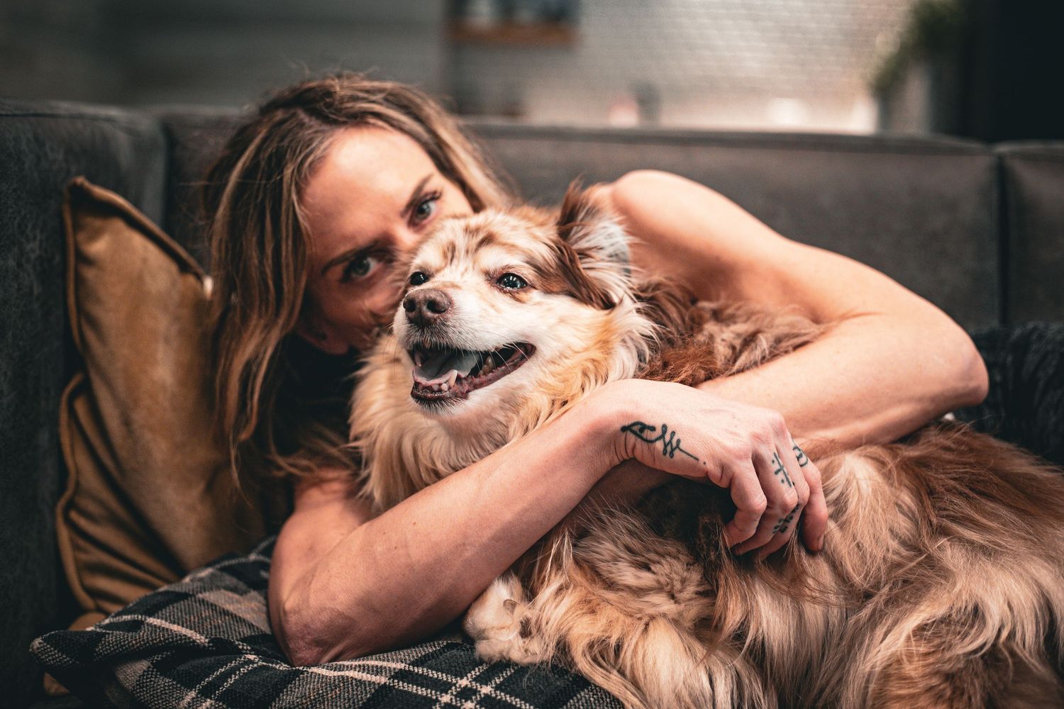 Woman with tattoos lovingly embracing her fluffy golden dog while relaxing on a gray sofa.