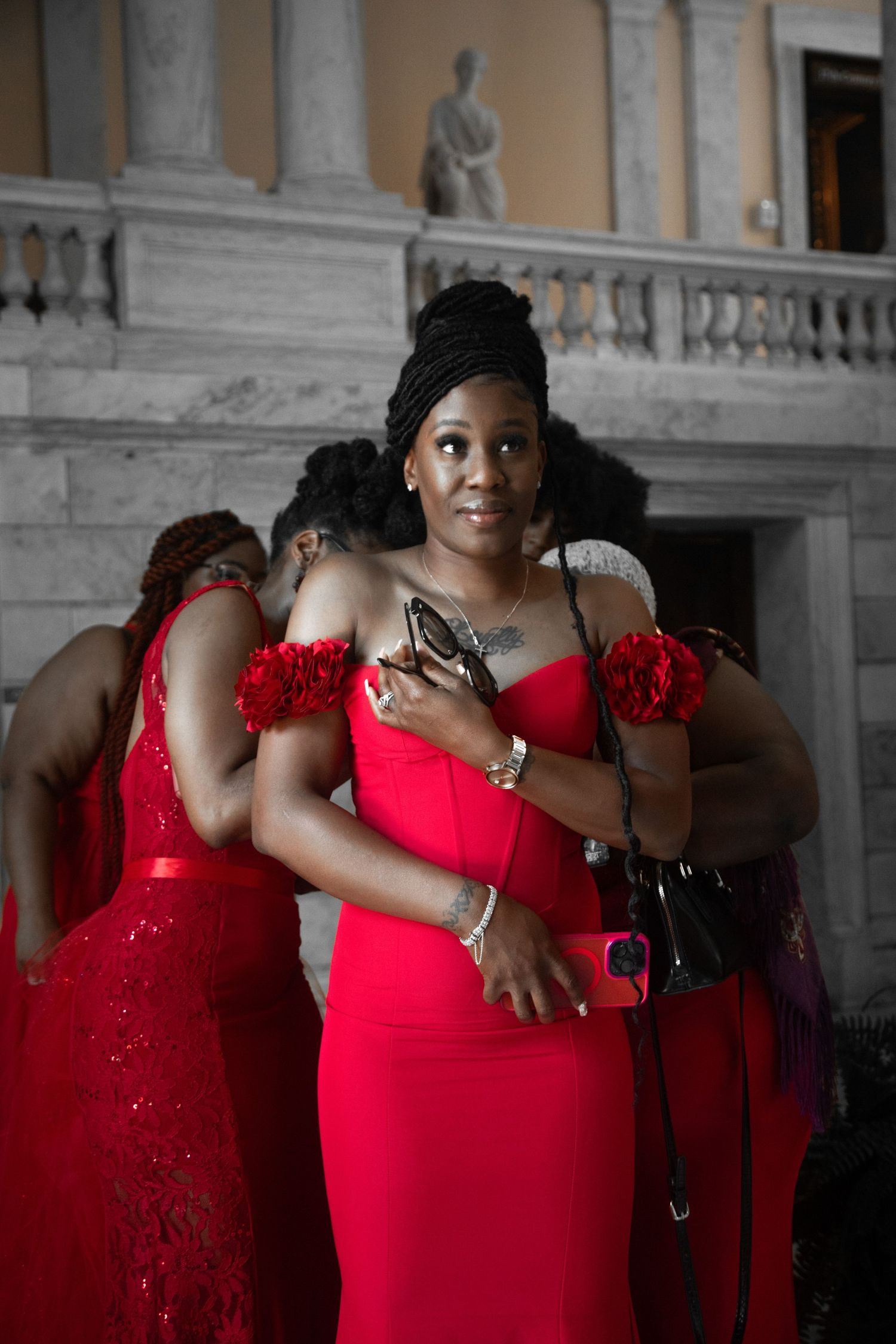 Elegant woman in red off-shoulder dress holds sunglasses indoors at a classical architectural venue with others nearby.