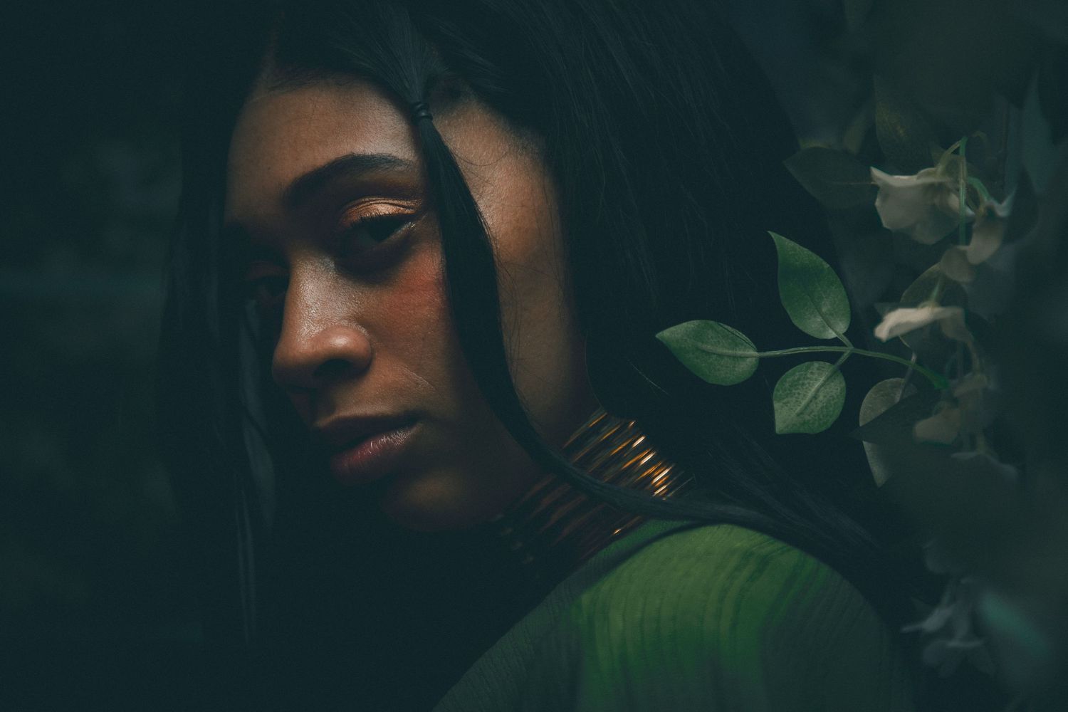 Moody portrait of woman with gold necklace beside white flowers in dark setting.