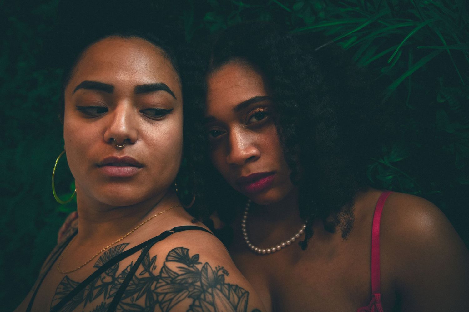 Two women with tattoos leaning together against a dark green leafy background in a close portrait.