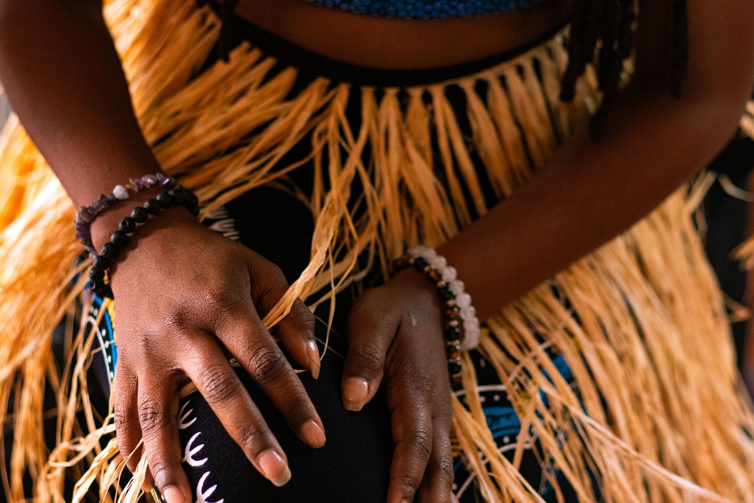 Close-up of hands adorned with beaded bracelets resting on traditional raffia grass skirt.