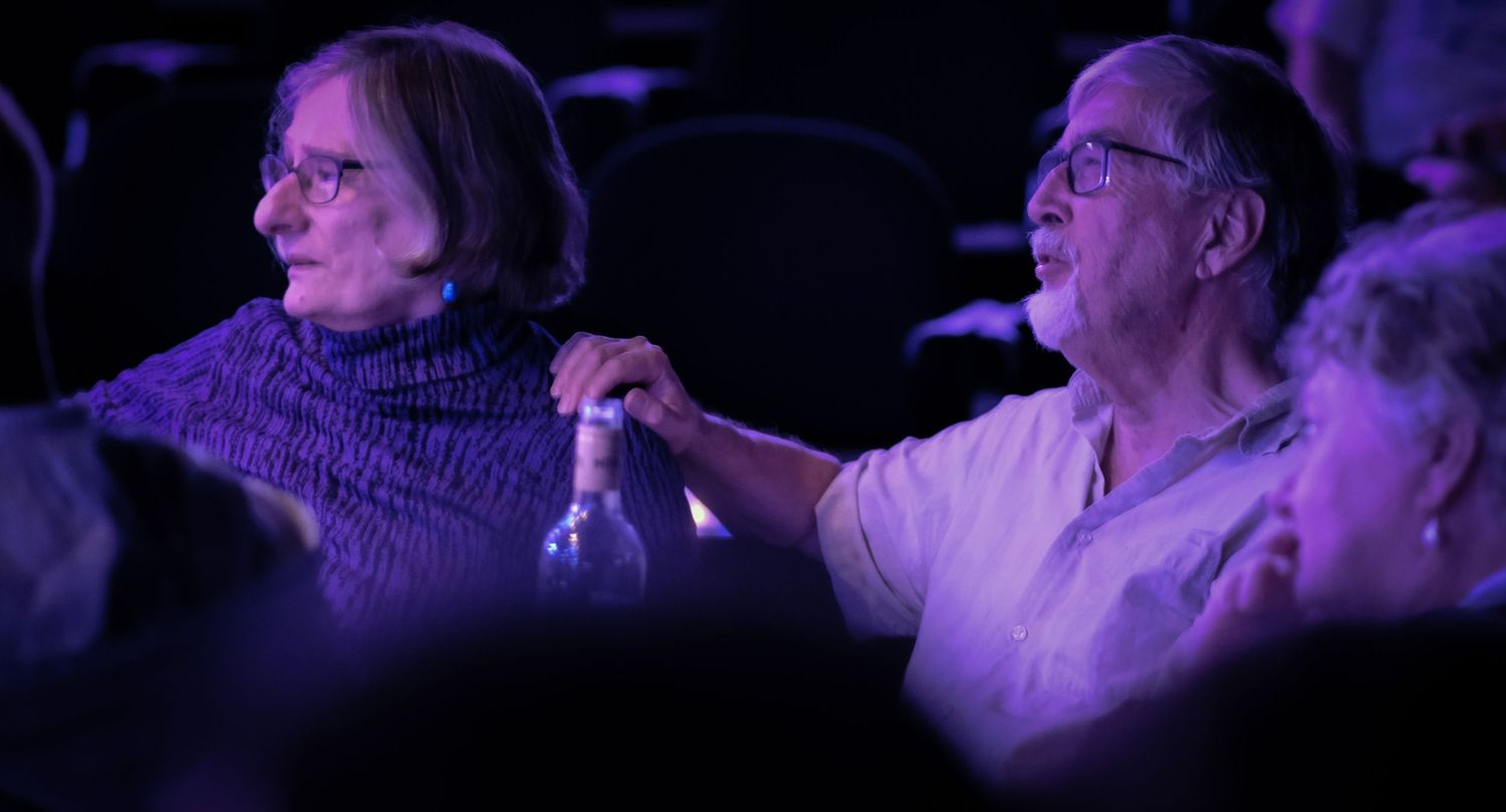 Elderly couple holding hands watching a performance in a purple-lit theater.