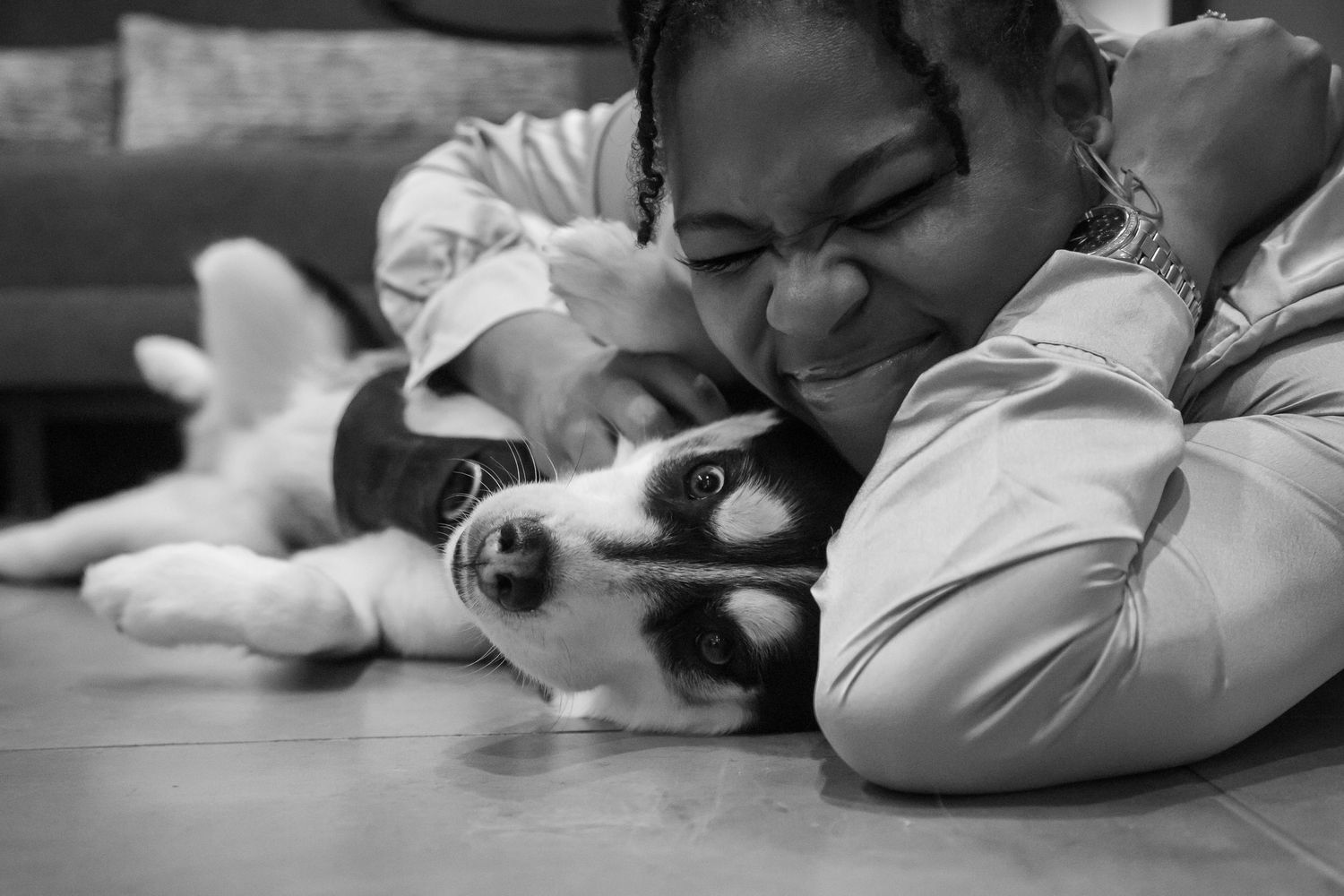 Black and white photo of a woman lying on the floor smiling while cuddling a Husky puppy closely.