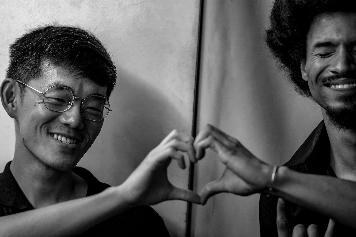 Two men smiling and making heart shapes with their hands in a black and white photo.
