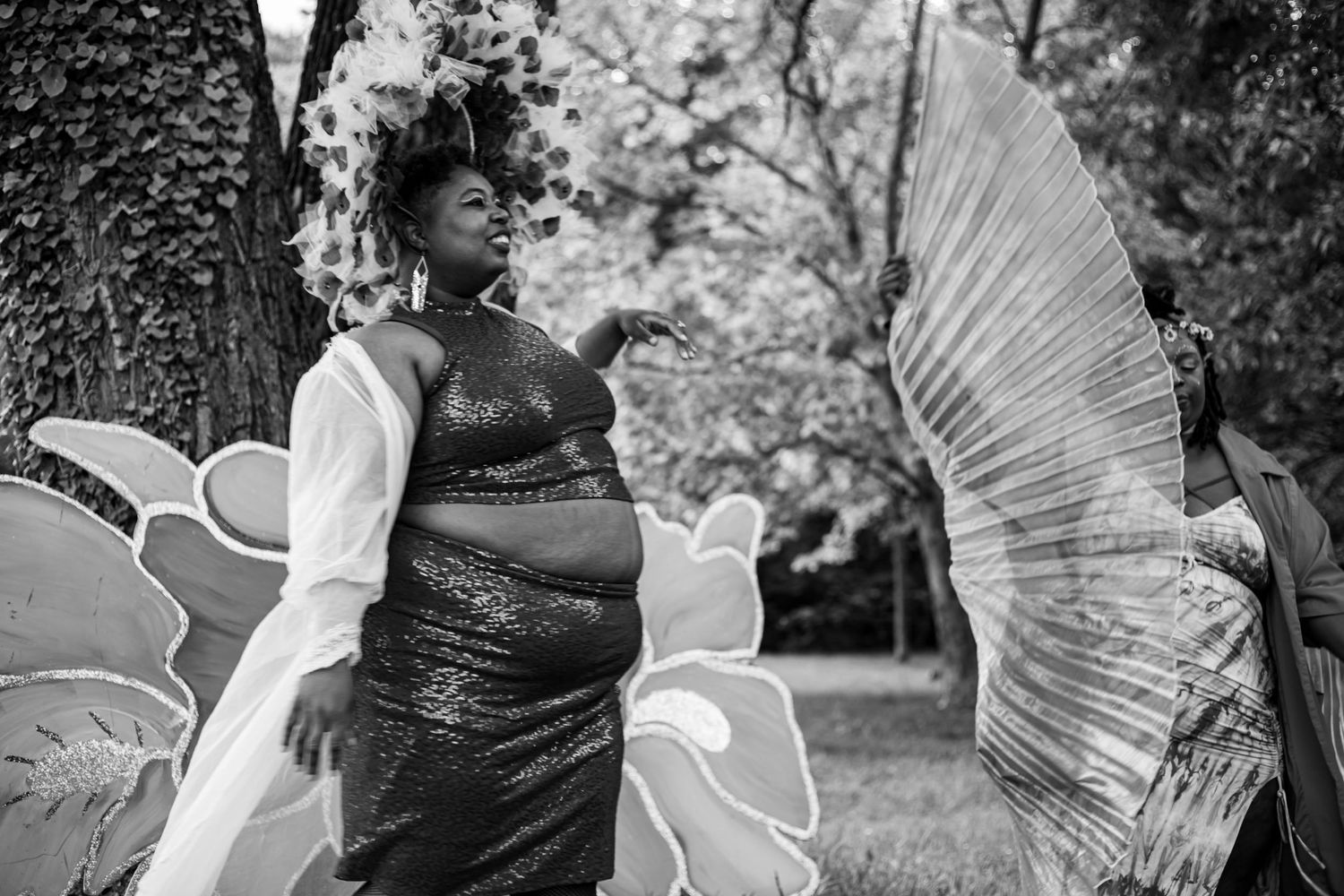 Black and white photo of a woman in a sequined dress posing outdoors with large decorative butterfly wings nearby.