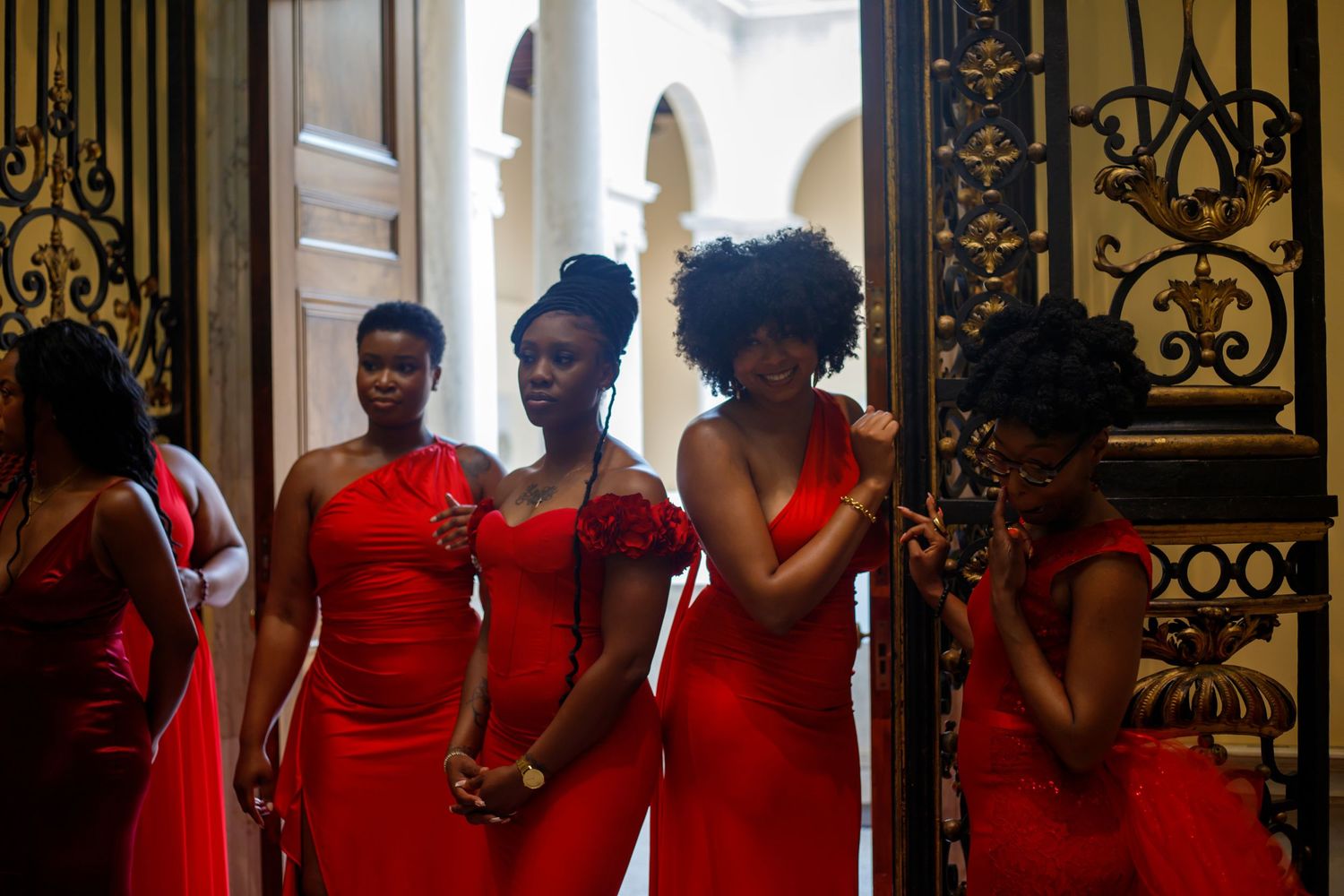 Four women in red formal dresses gathered near ornate black and gold iron gate.