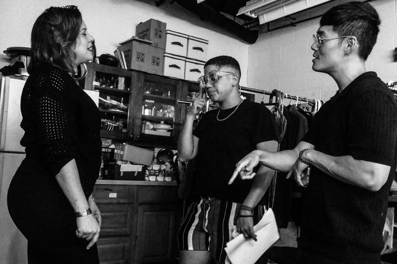 Black and white photo of three people in discussion inside a backstage storage room with shelves.