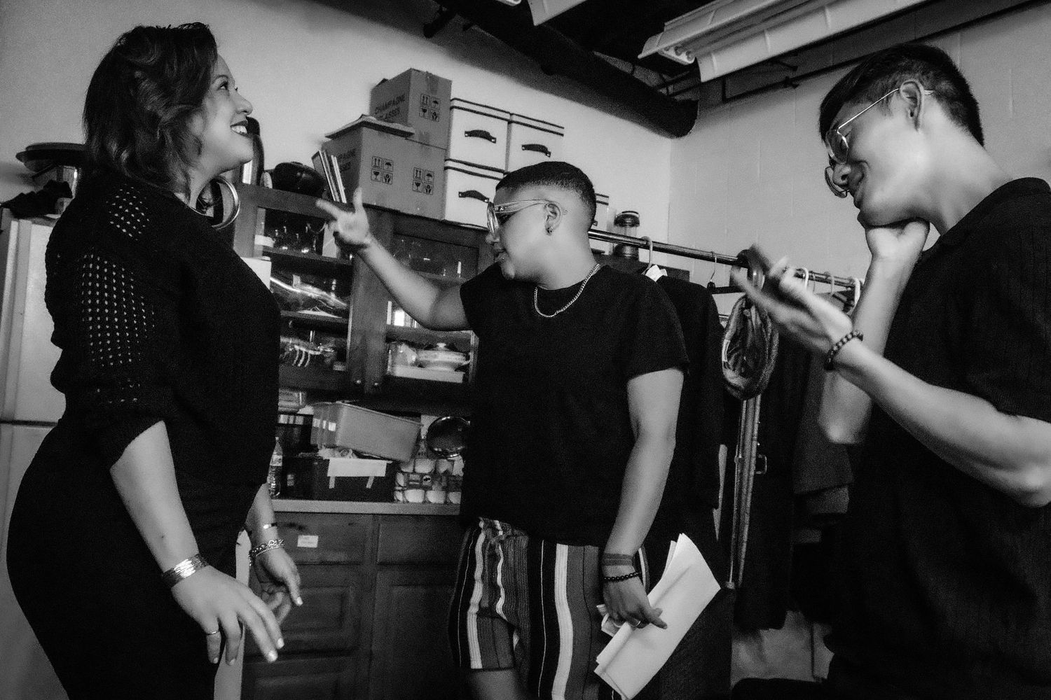 Black and white photo of three people having a conversation in a cluttered room.