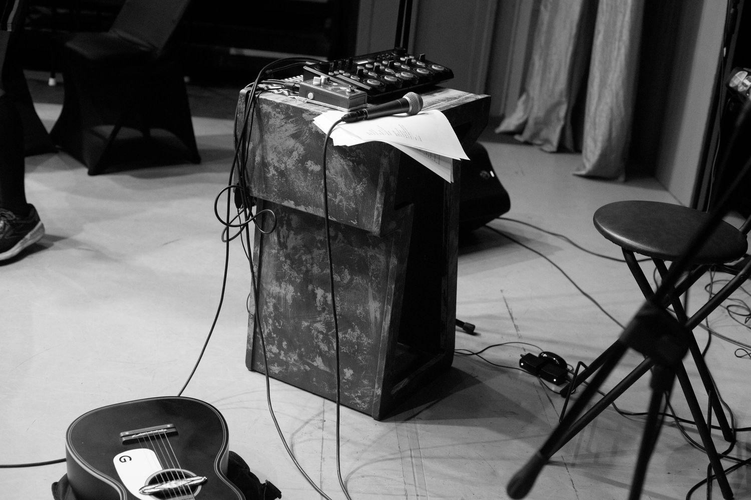 Black and white photo of music equipment including a guitar amp and pedals on a stage floor.