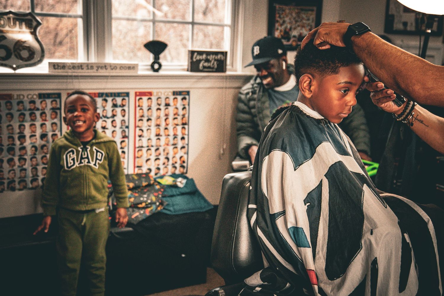Young boy smiling in a barbershop while another child gets a haircut from a skilled barber.