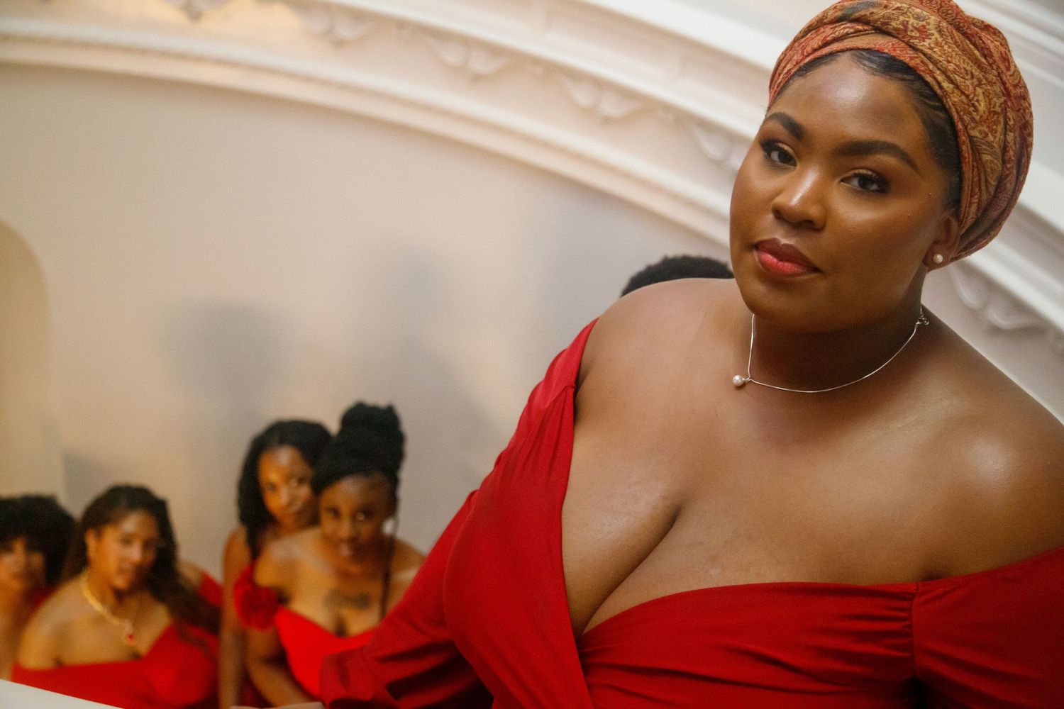 A Black woman in a red off-shoulder dress and orange headwrap poses confidently with bridesmaids in red behind her.