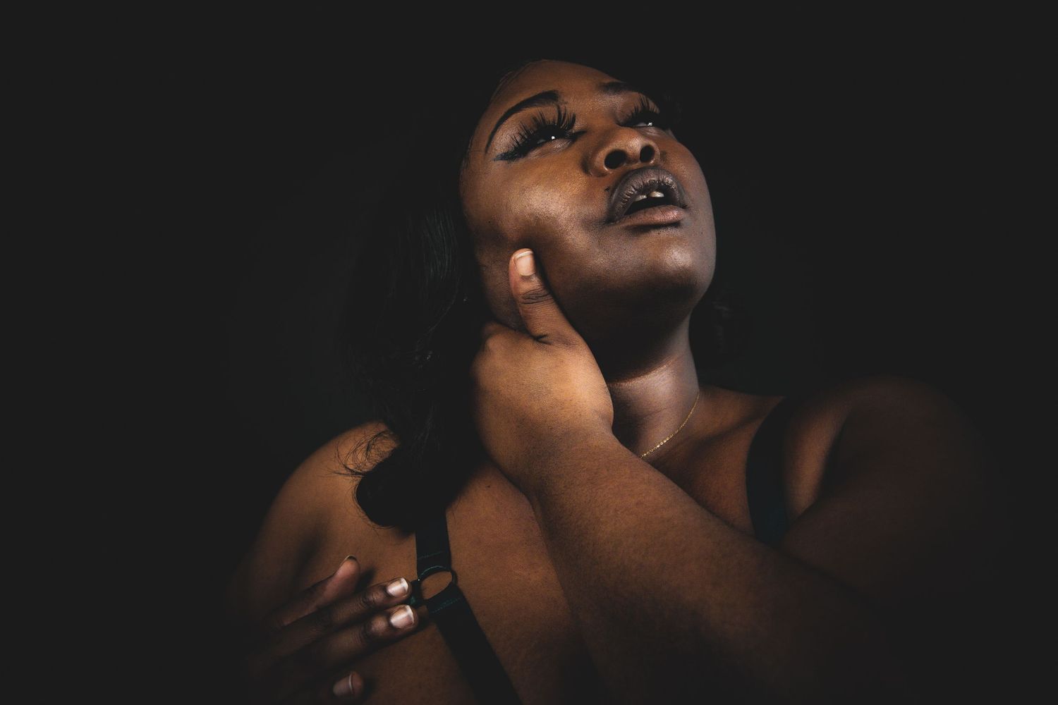 A Black woman in a dramatic low-key portrait, hand raised to her cheek, gazing upward in dark lighting.
