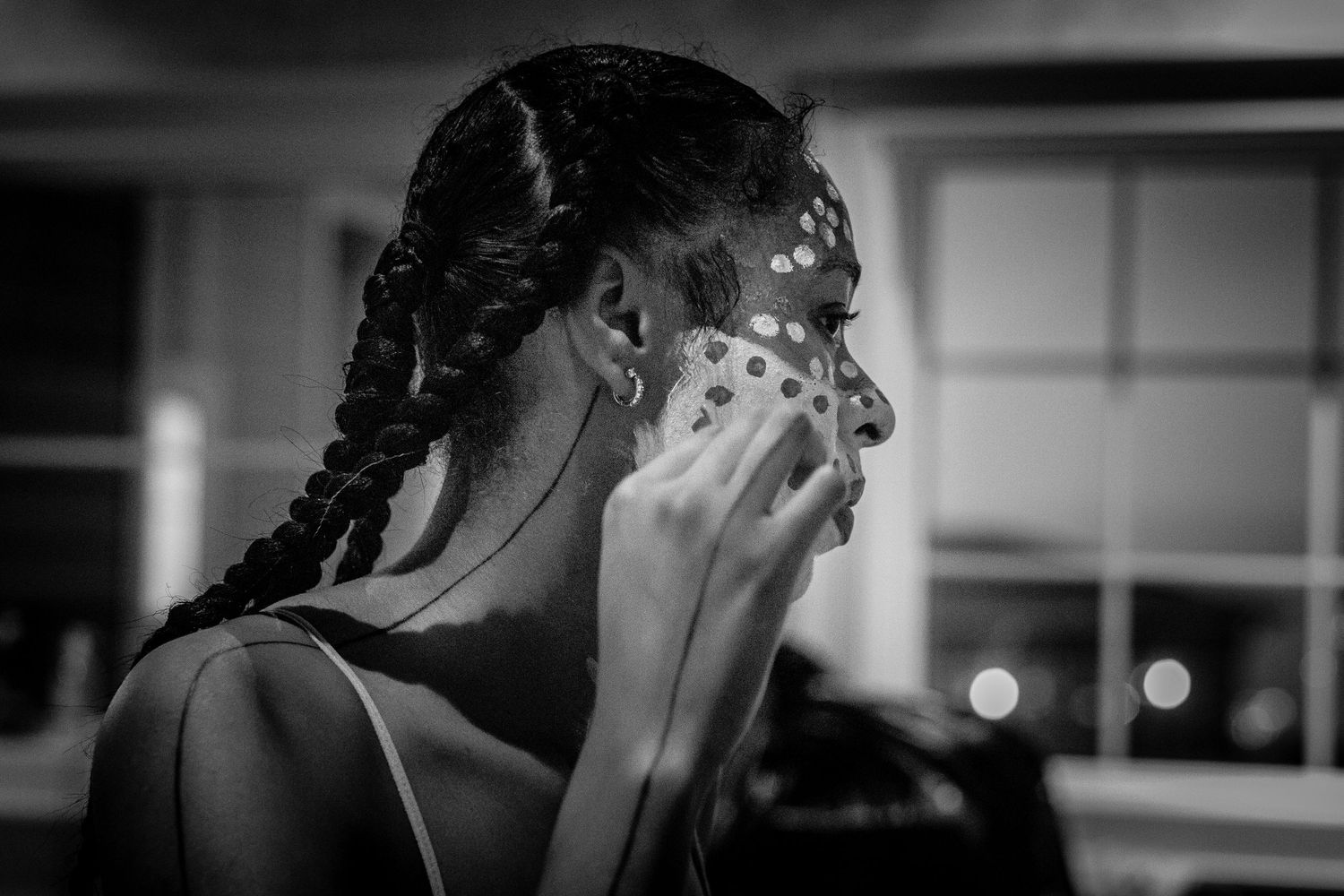 Black and white image of a woman with braids applying white dot face paint to her own cheek.