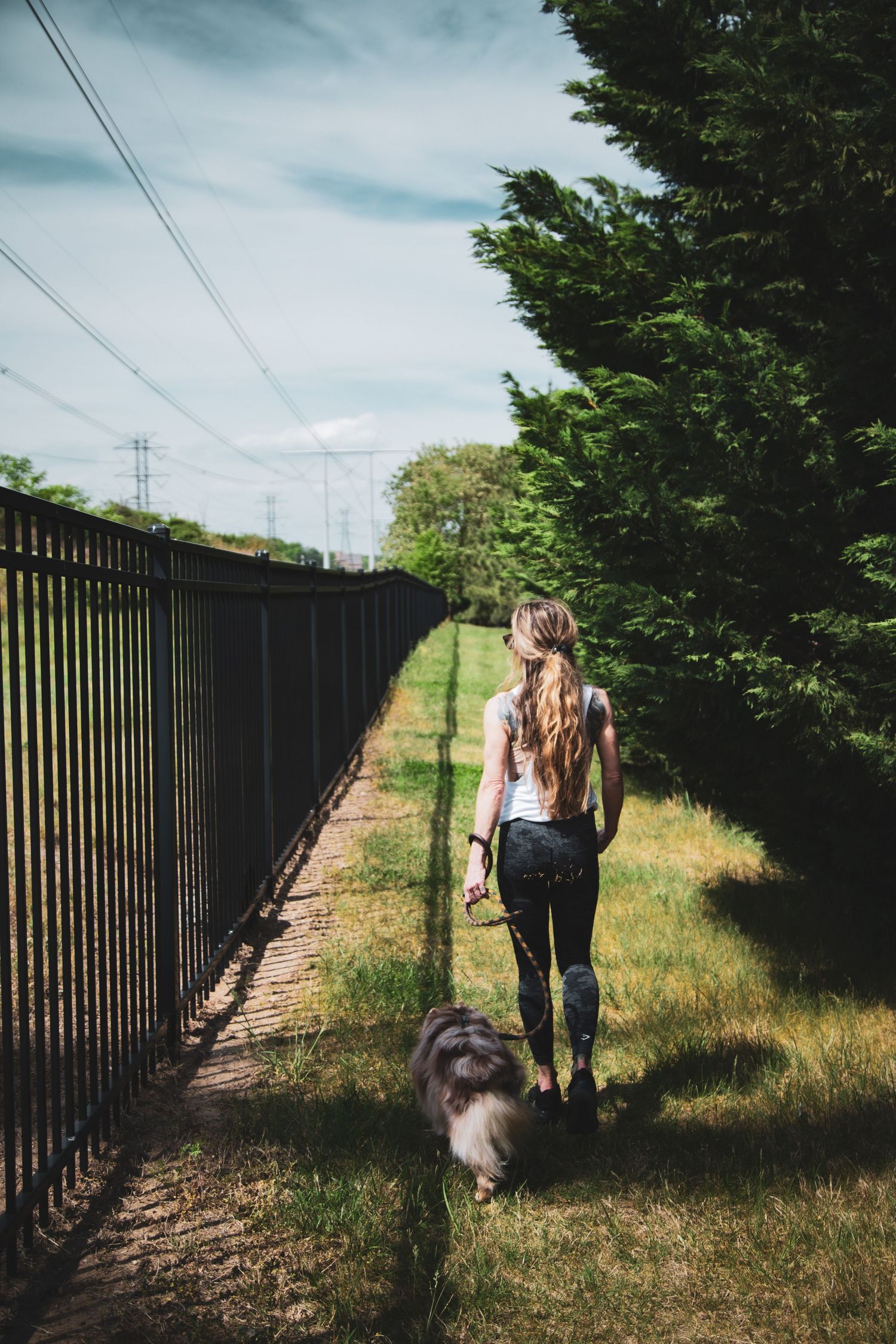 Woman walking her fluffy Australian-shepherd dog along a fenced path surrounded by green trees.