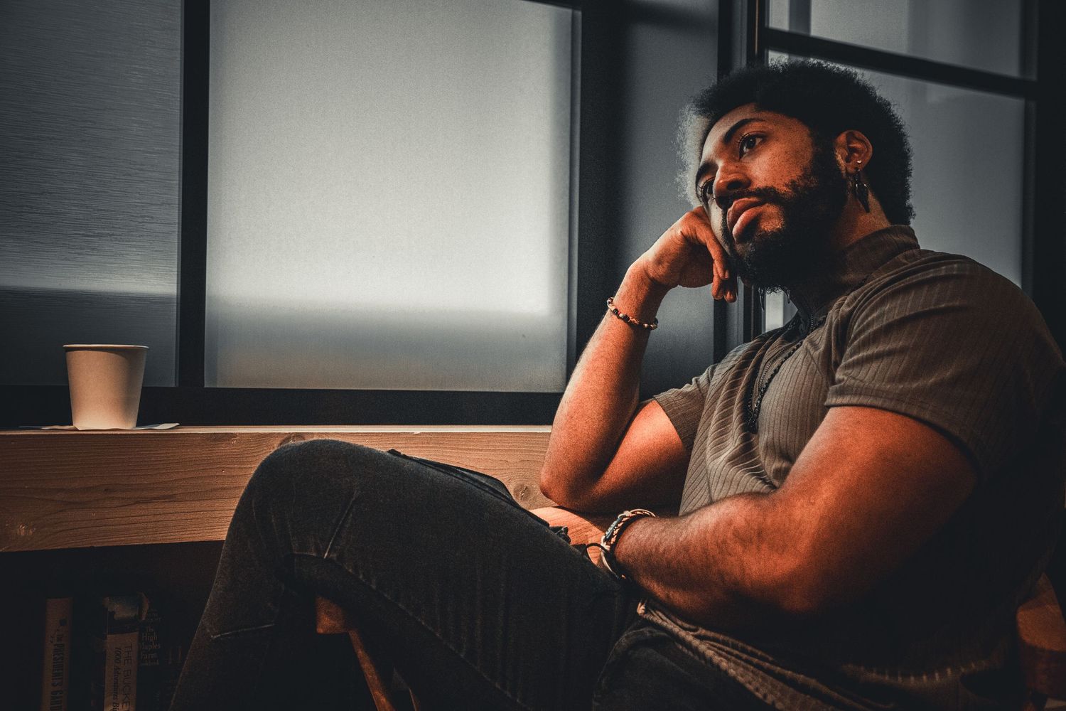 Bearded man sitting thoughtfully by a window in a dimly lit room.