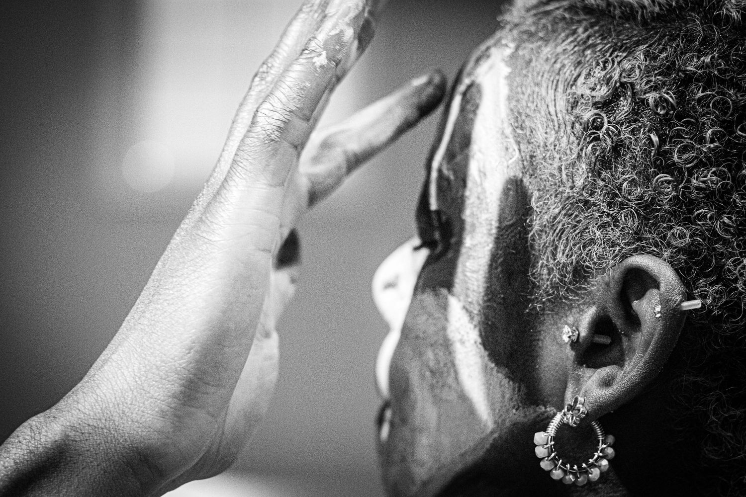 Black and white close-up of a hand applying white face paint to a performer wearing decorative earrings.