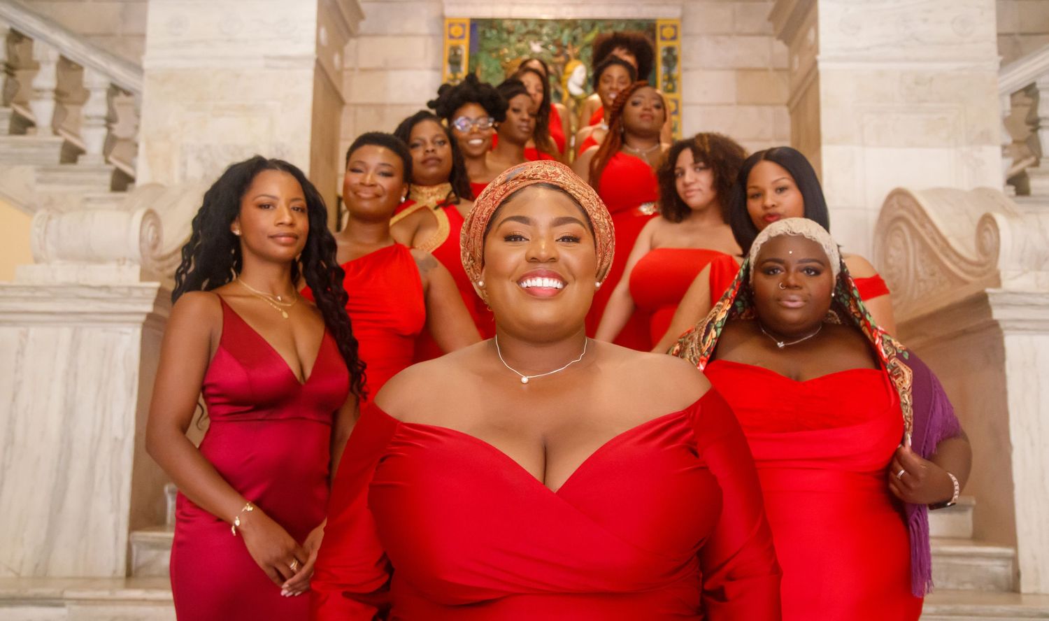 Group of black women who look like Bridesmaids in vibrant red dresses posing on a luxurious marble staircase.