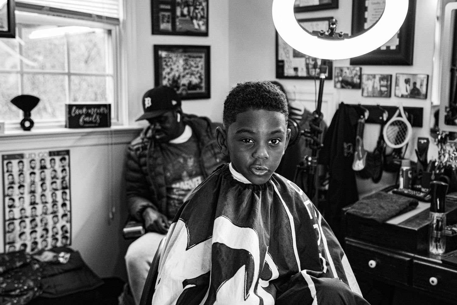 Black and white portrait of a young boy in a barber cape sitting in a barbershop chair.