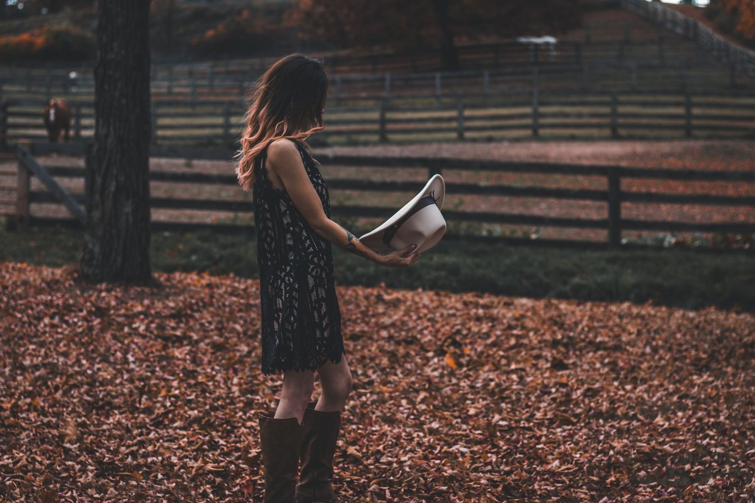 Young woman in floral dress reading a book in a rustic farm setting covered in fallen leaves.