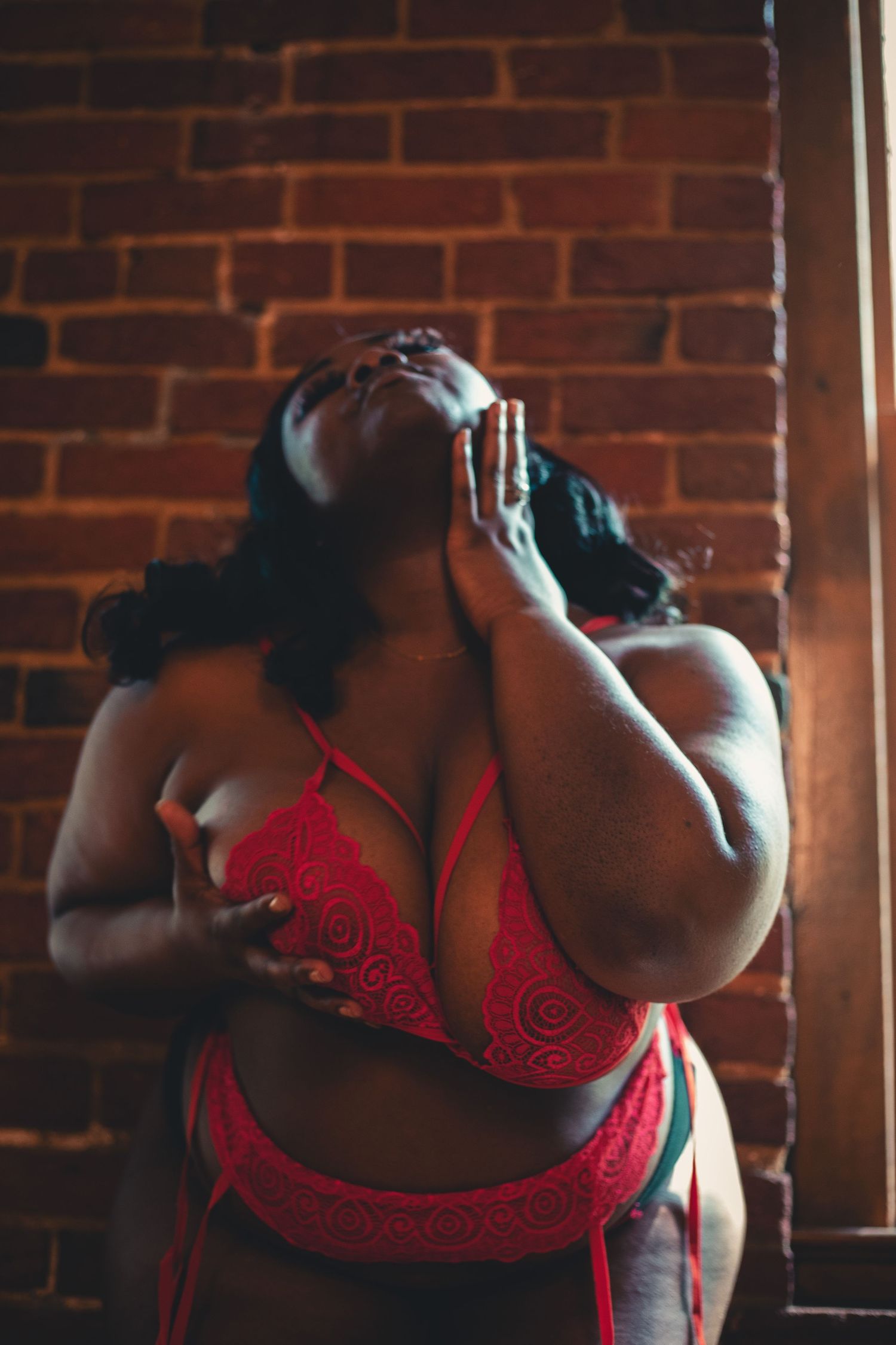 Plus-size woman in red lace lingerie posing dramatically against a rustic brick wall in moody lighting.