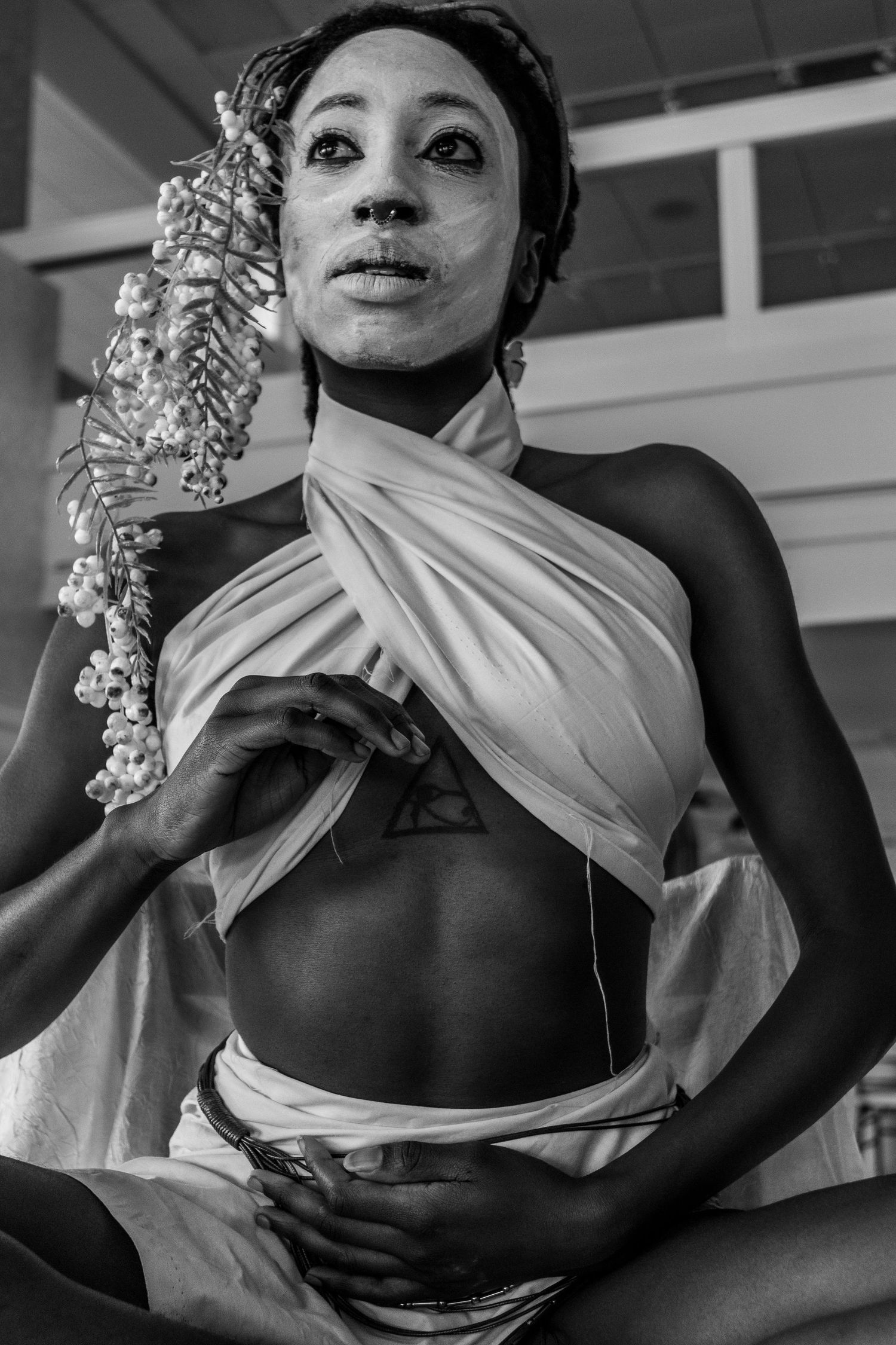 Black and white photo of a model with white face paint wearing a draped white fabric costume with floral hair.