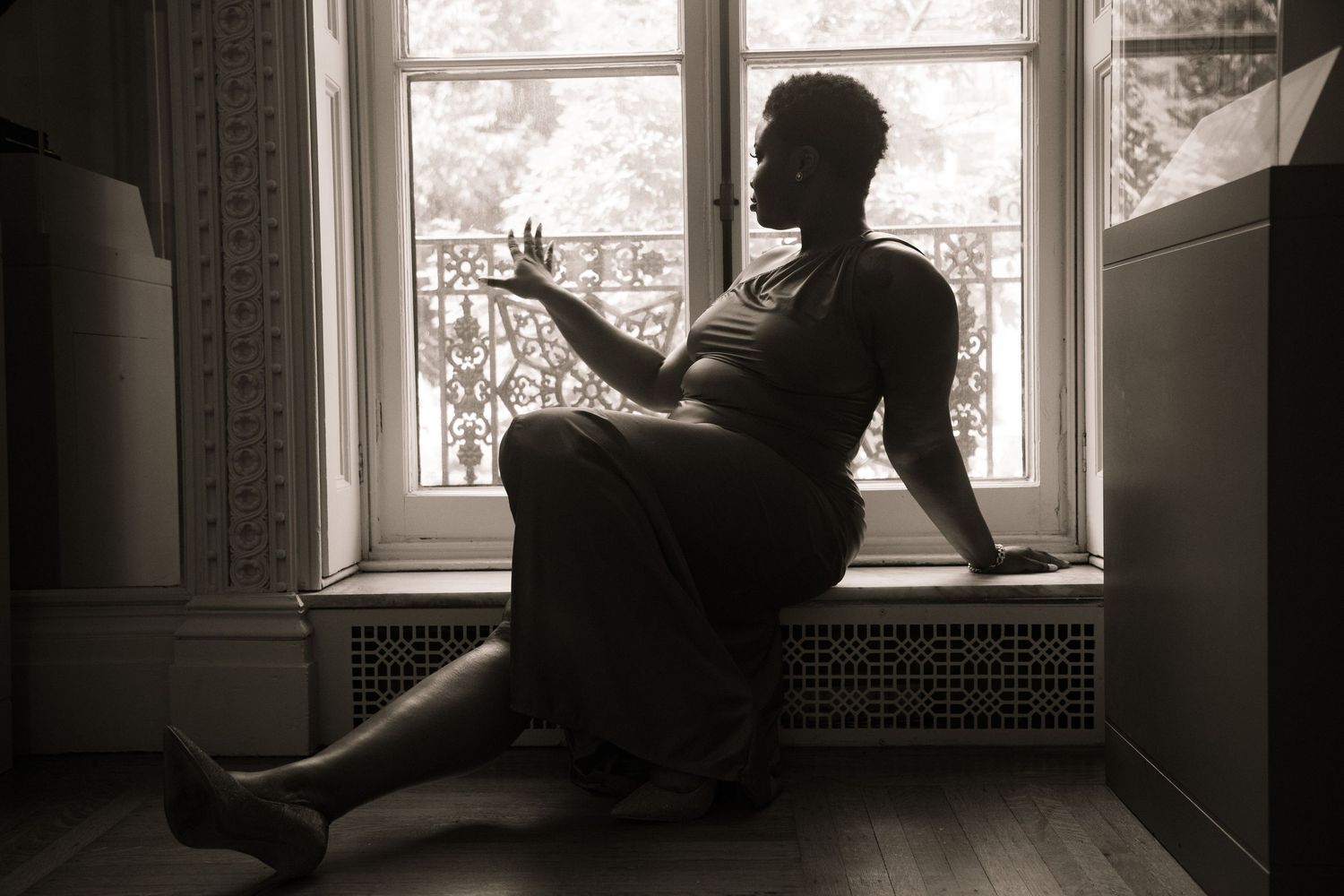Black and white silhouette of a woman sitting by a vintage window, reaching toward the glass.