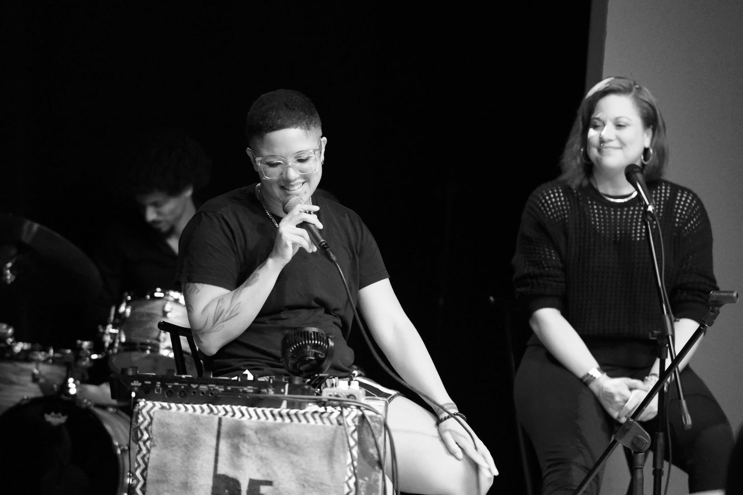 Black and white photo of two women on stage, one speaking into a microphone while the other smiles beside her.