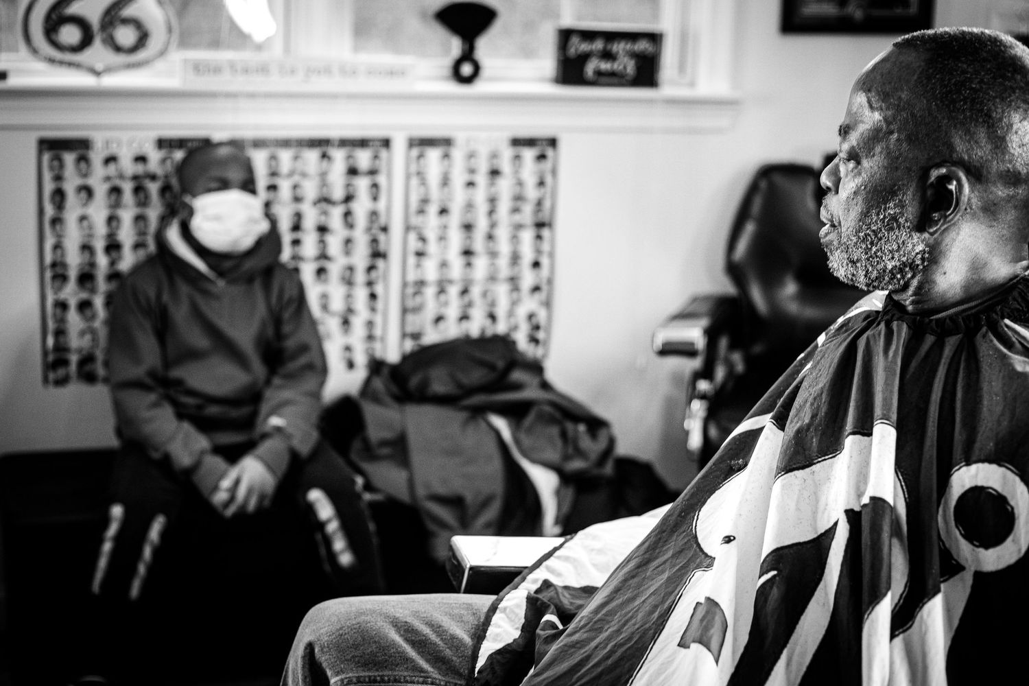 Black and white image of a barbershop client sitting while a masked person waits in the background.