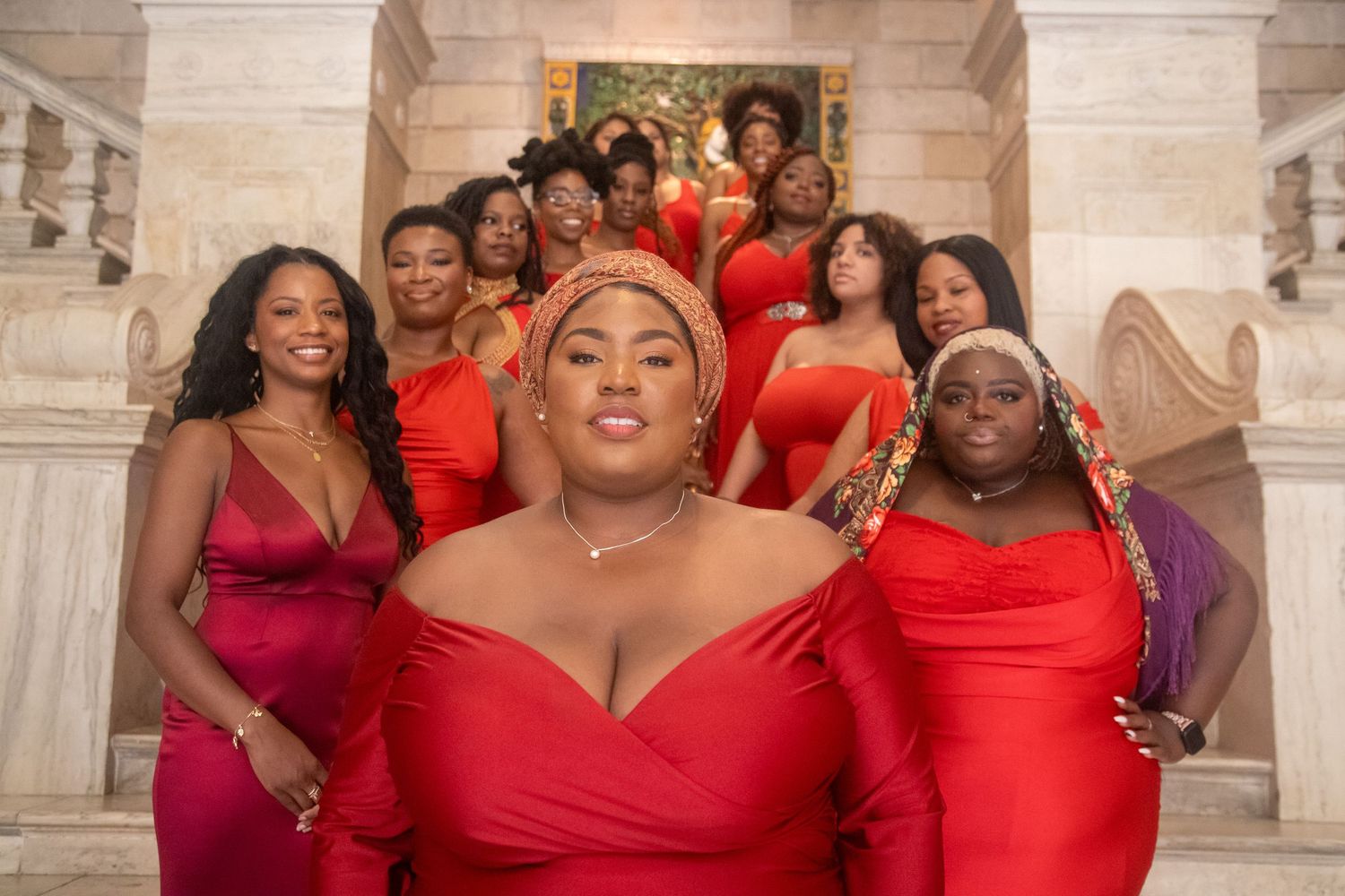 Group of black women in red dresses posing on marble staircase for elegant formal photoshoot.