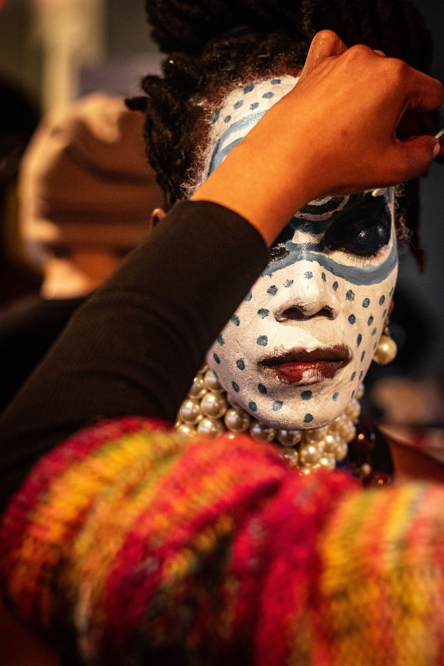 Close-up of white face paint with blue dots being applied to a performer wearing pearl necklaces.